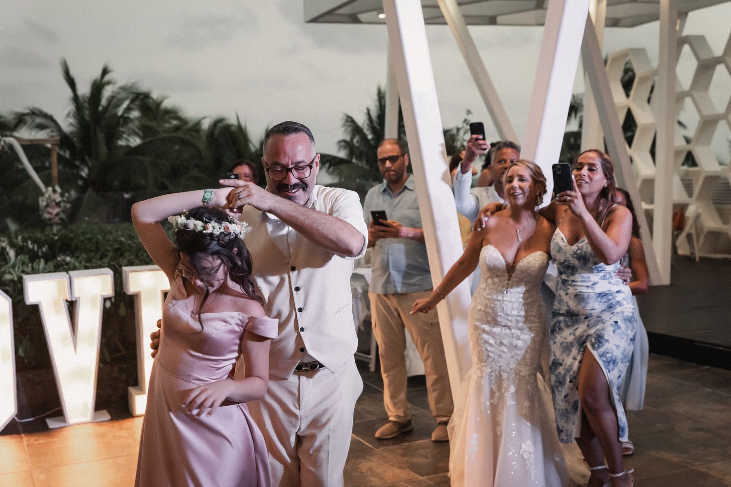 Groom places flower crown on bride during lively destination wedding reception celebration with guests.