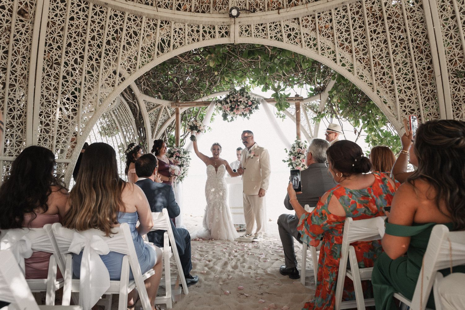 Newlyweds celebrate recessional under ornate white arch as guests cheer at outdoor beach wedding.