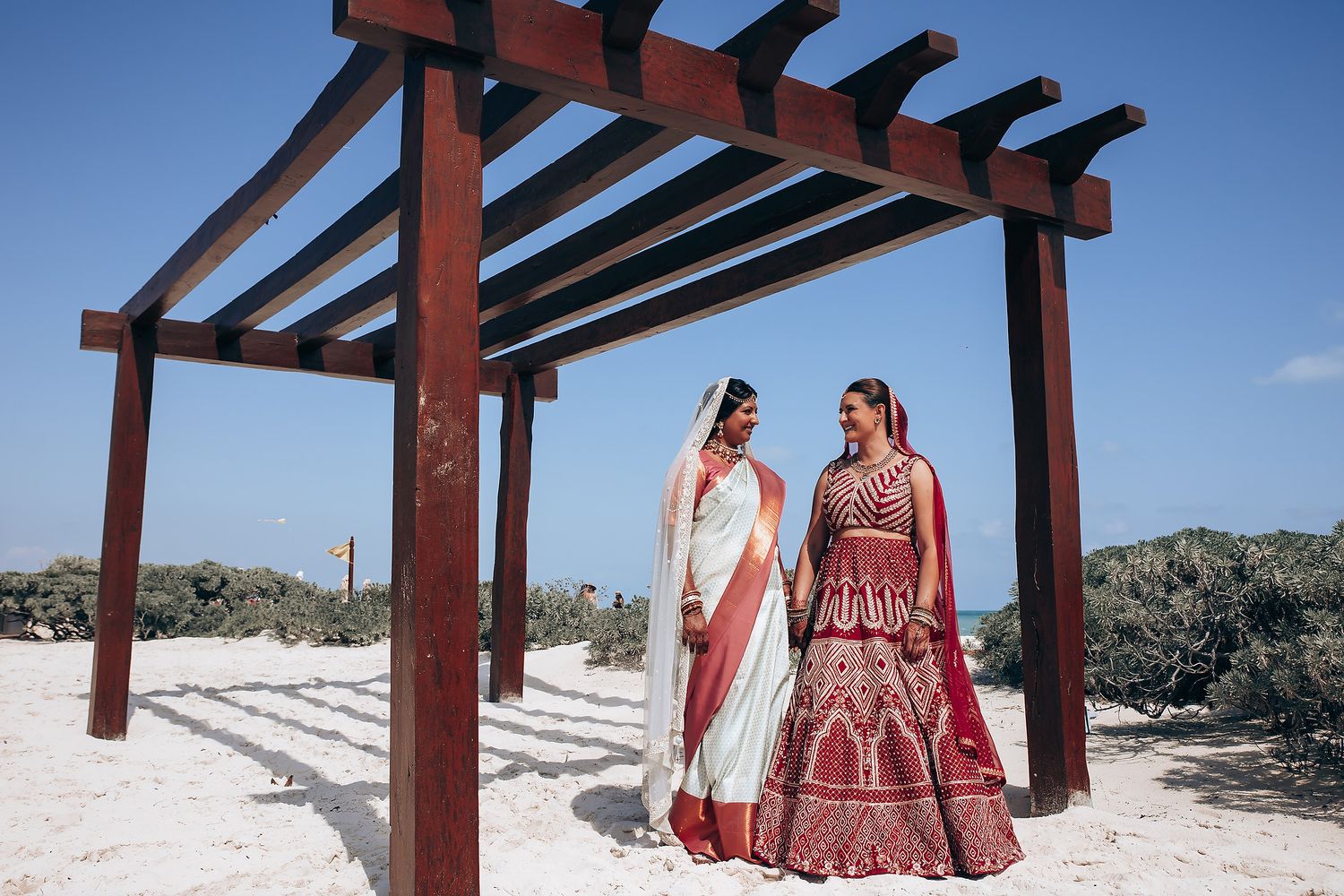 Two people in traditional Indian wedding attire pose under a wooden pergola against a bright blue sky.