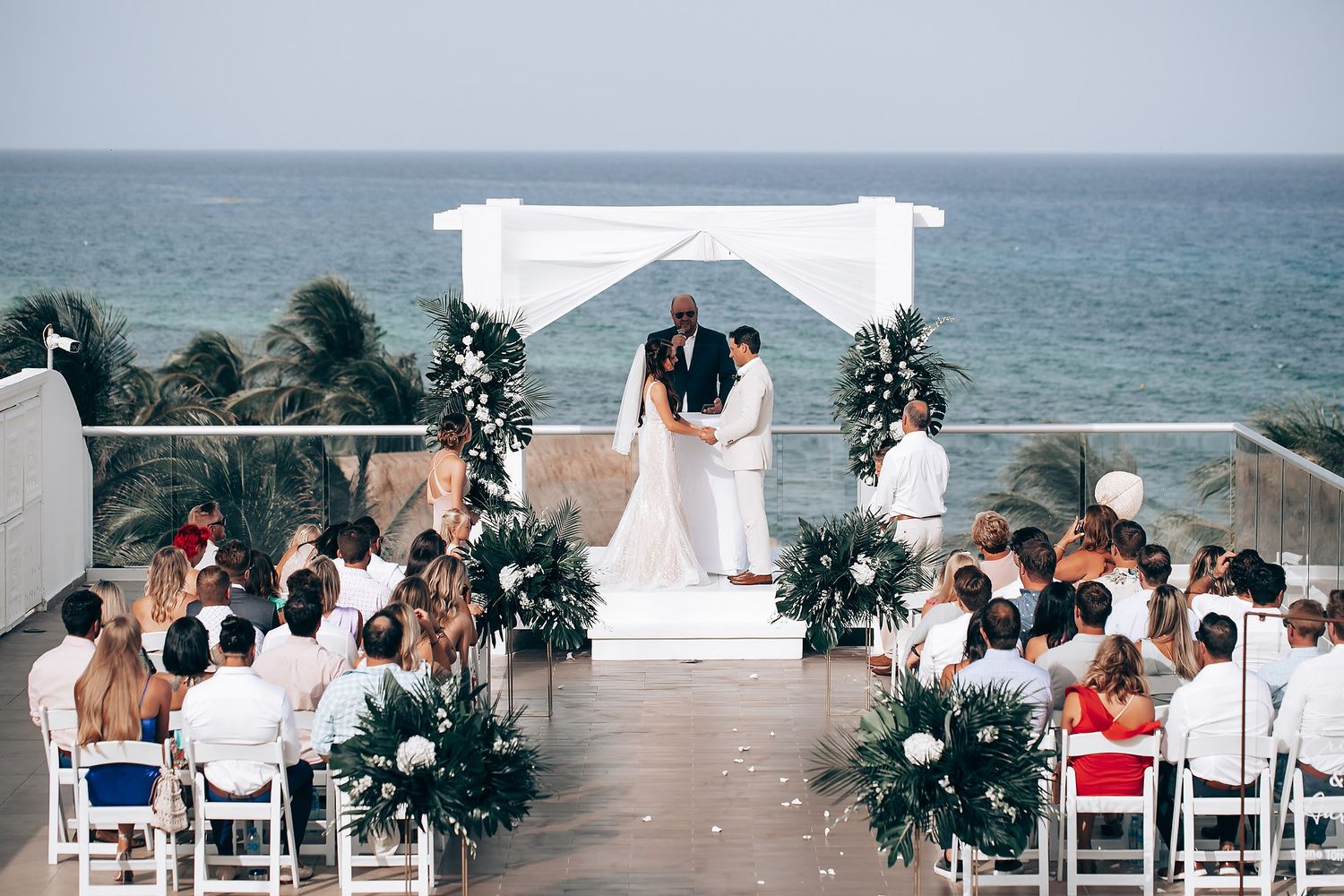 Panoramic view of a beach wedding ceremony setup with white chairs and tropical decor overlooking the ocean.