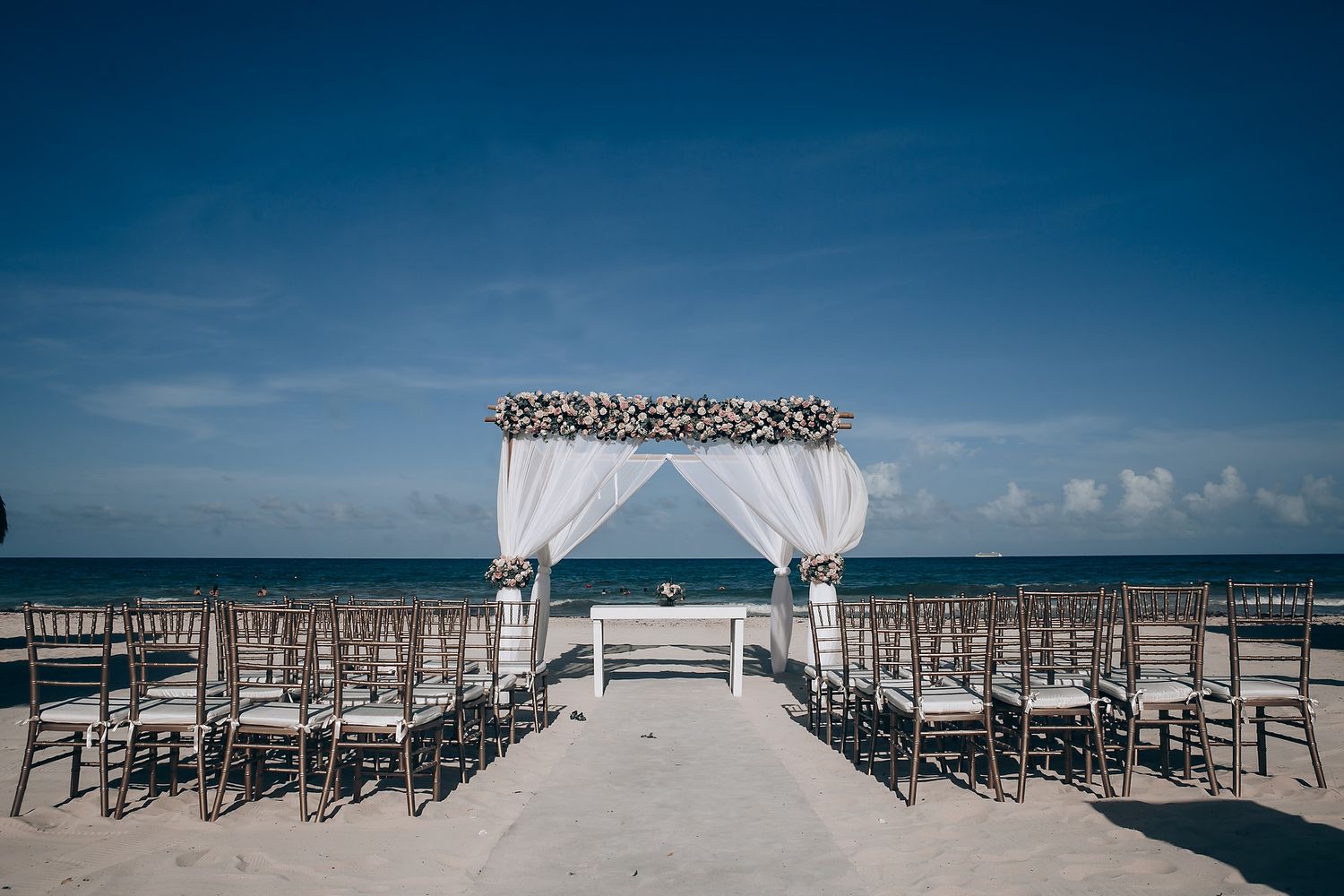 Beachfront wedding ceremony setup with white chairs and floral arch against blue ocean backdrop.