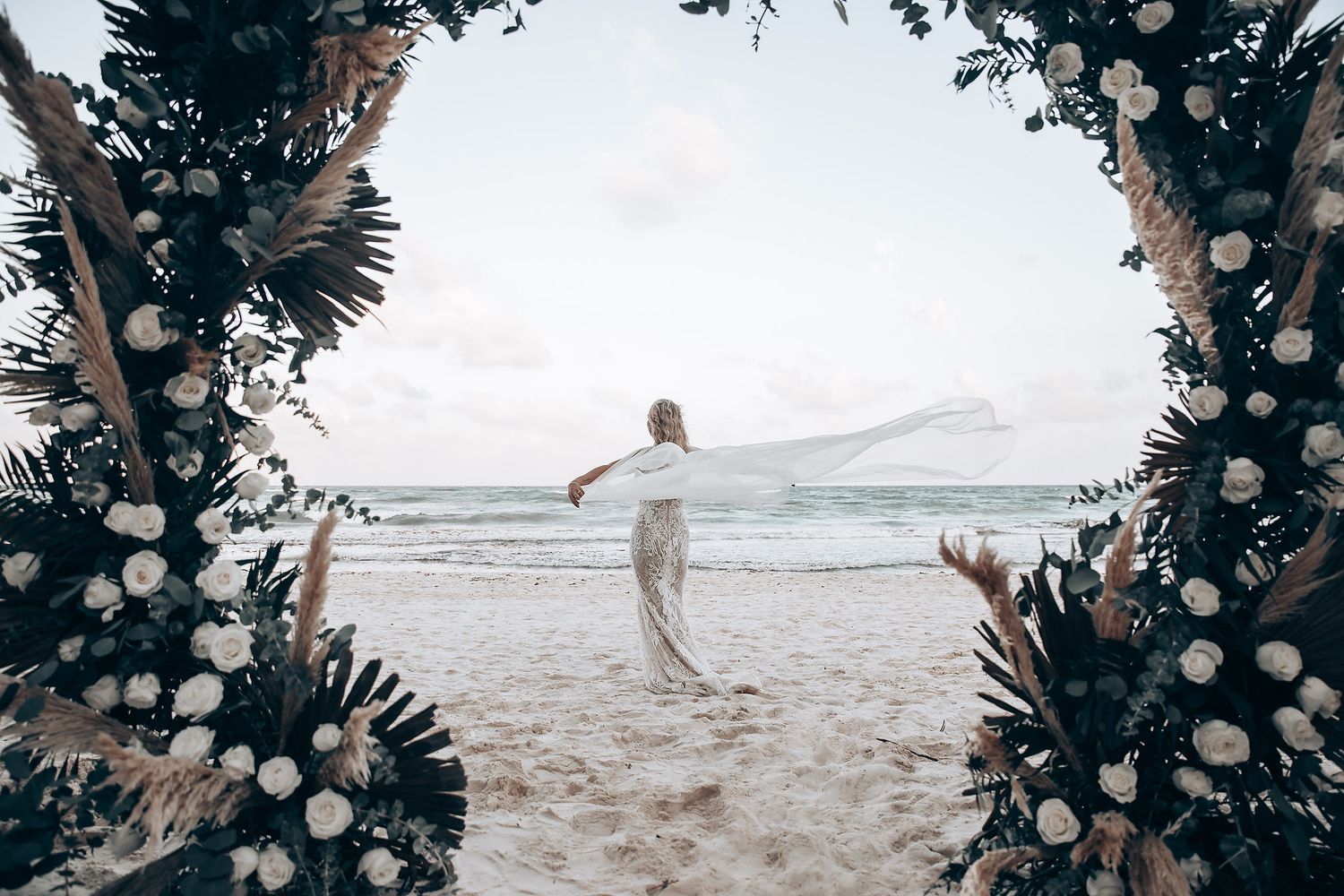 A person in white walks on a beach framed by a floral arch with ocean views.