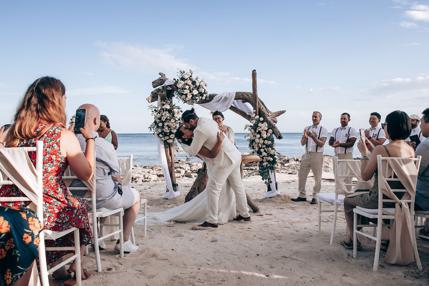 Beach wedding ceremony in progress with guests seated on white chairs by the ocean.