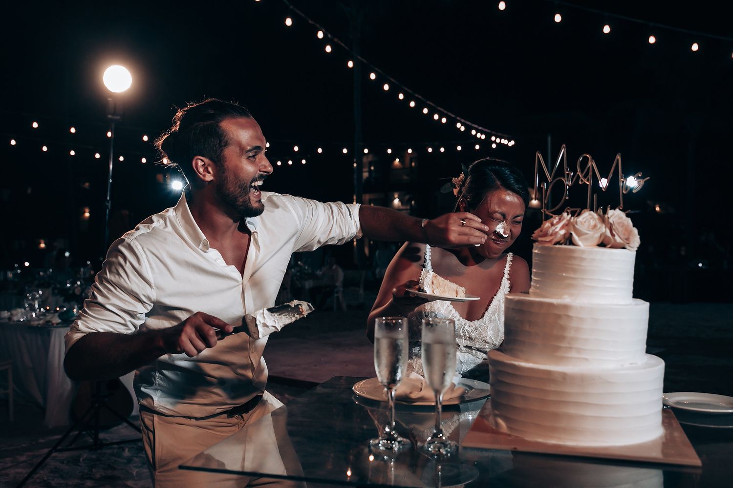 A wedding couple cutting their multi-tiered cake under string lights at night.