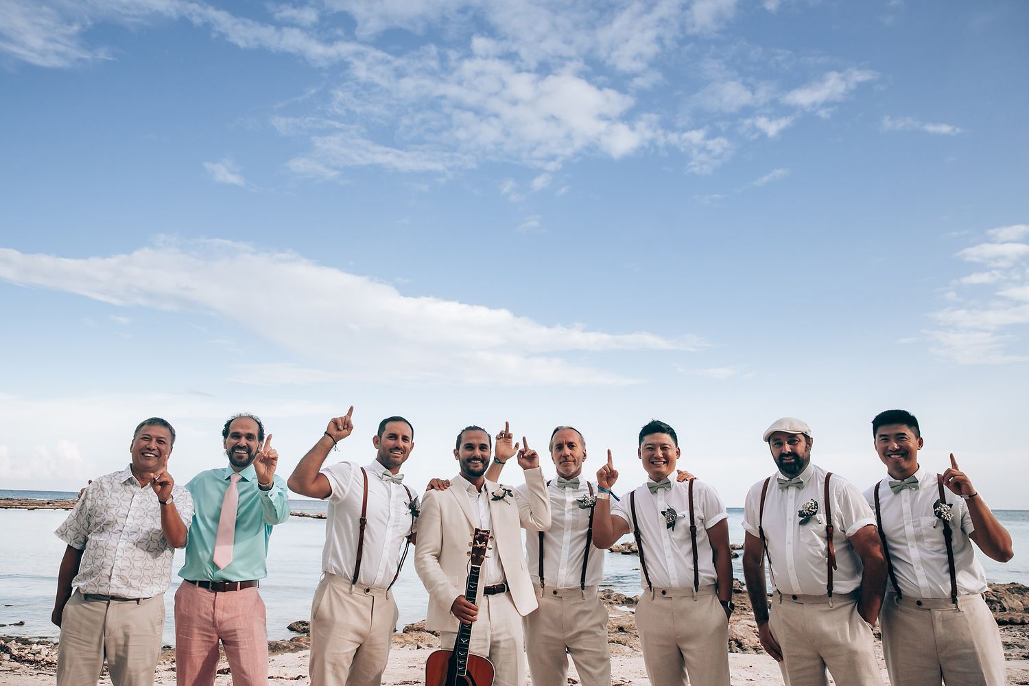 A group wearing light colored attire poses together on a sunny beach with blue sky and clouds in the background.