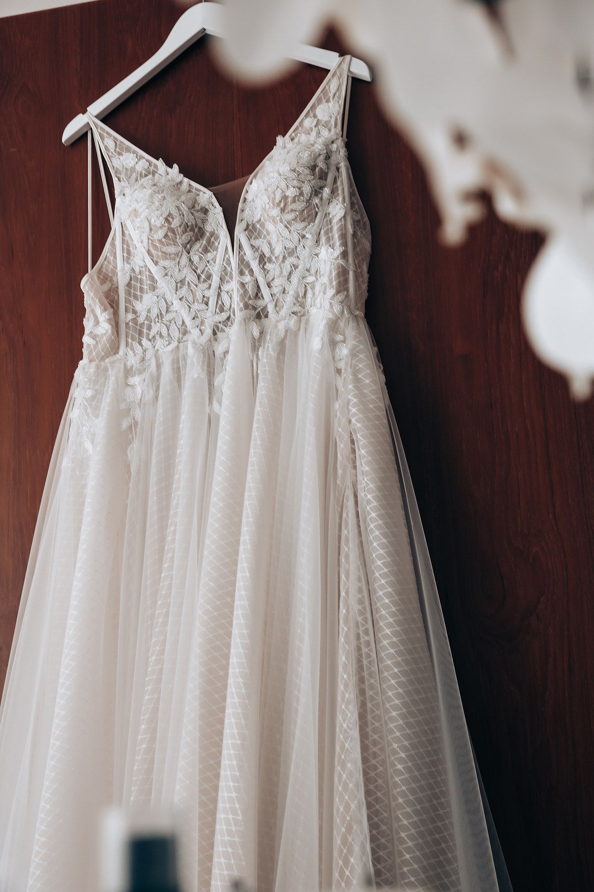 Delicate white lace wedding dress with floral details hanging against dark wooden background.