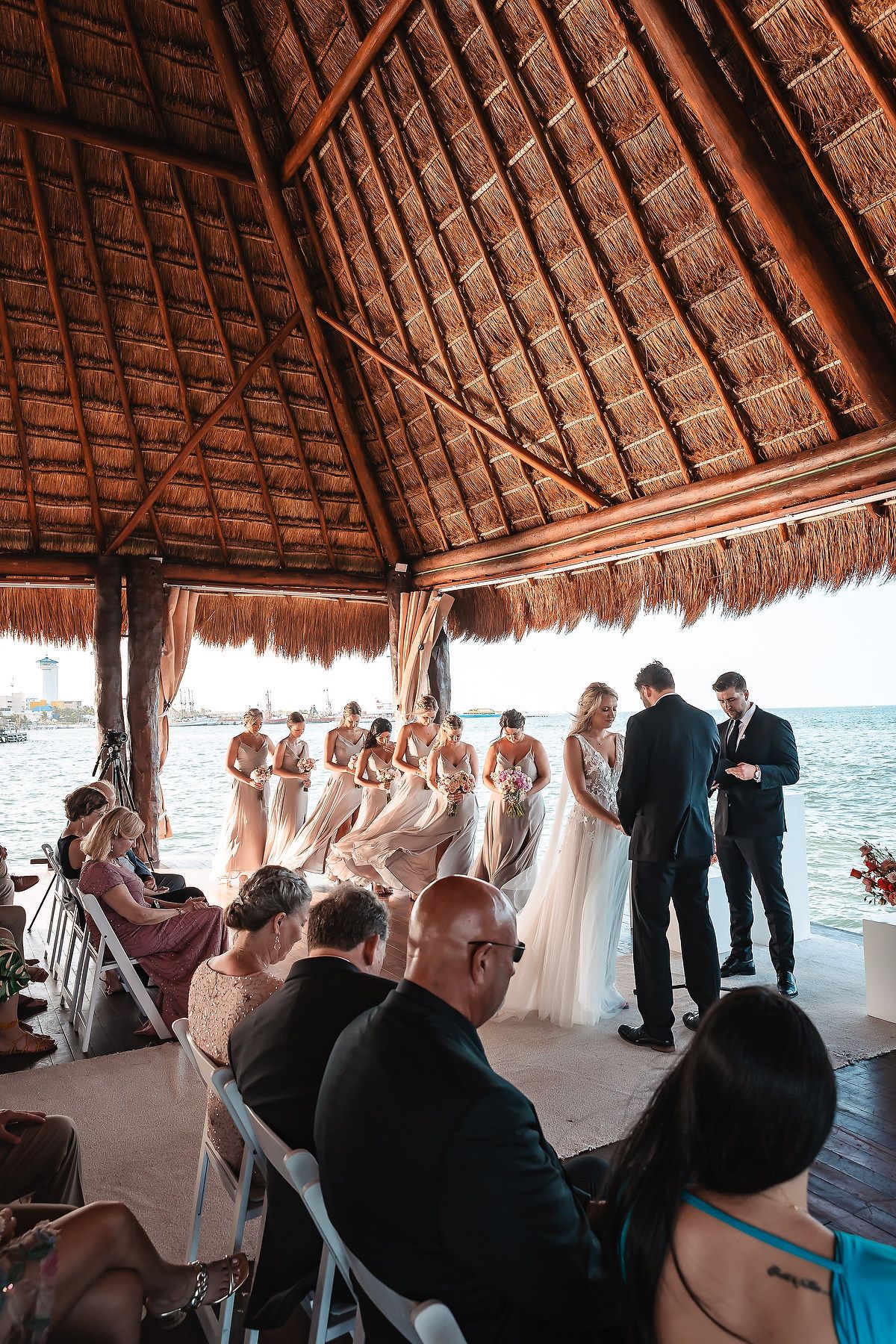 Wedding ceremony in progress under a rustic thatched palapa overlooking the beach.