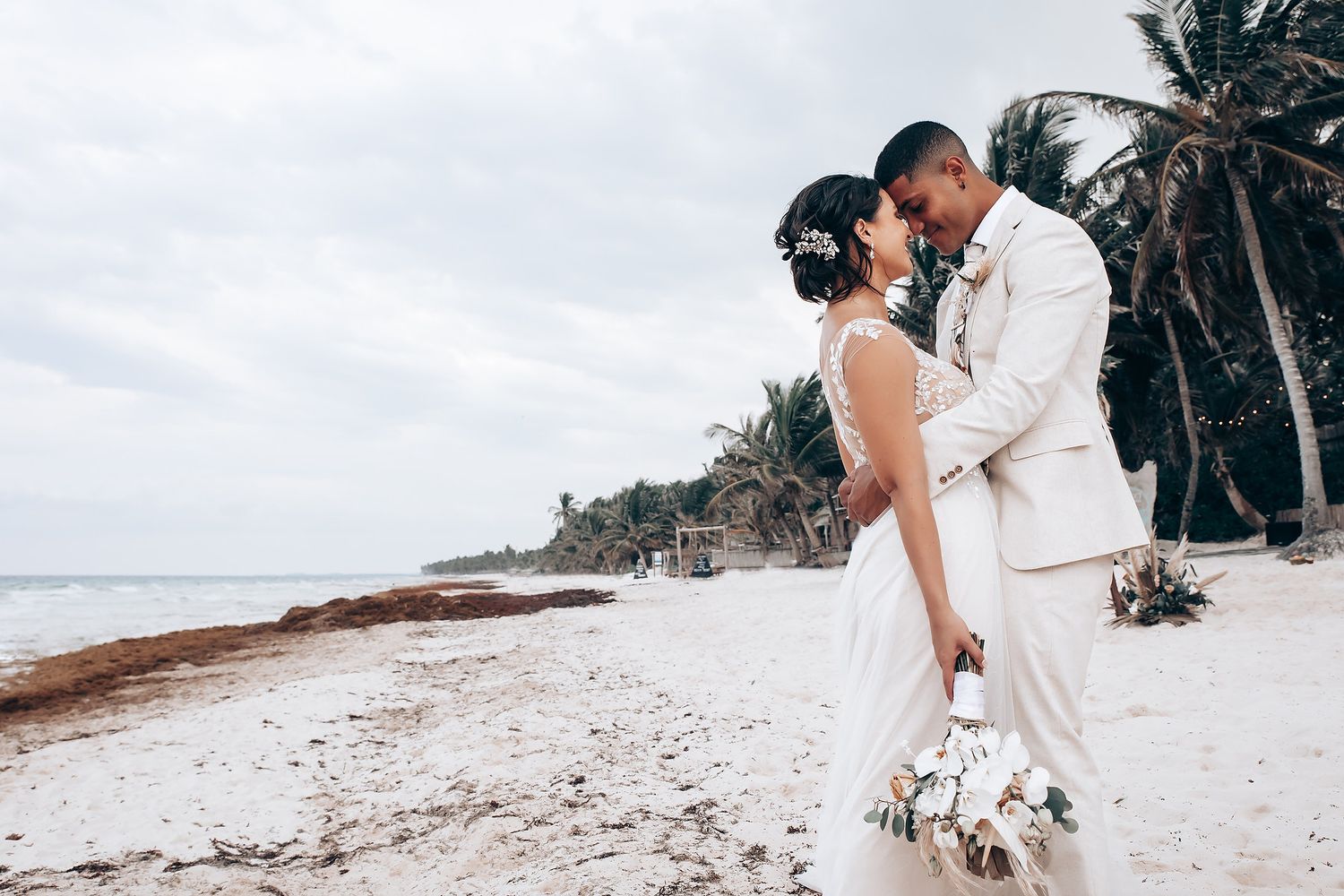 Romantic beach wedding ceremony setup with white decor and palm trees along the shoreline.