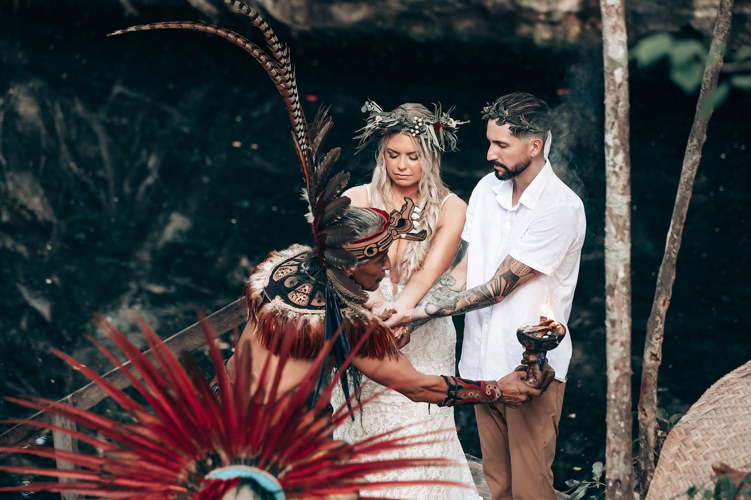 A couple participates in a traditional ceremony with native American feather decorations in a forest setting.