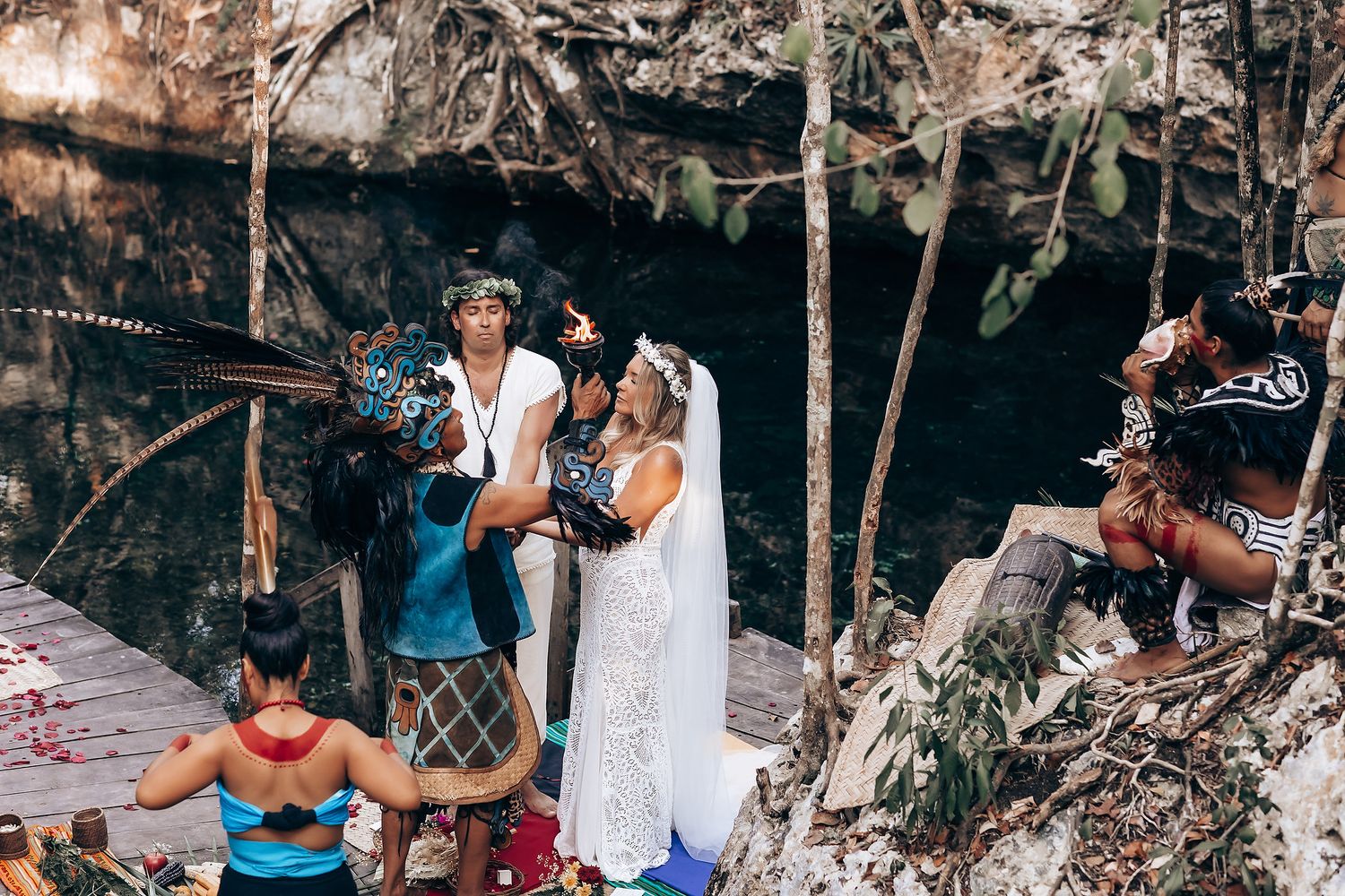 People gather in a shaded outdoor area surrounded by trees and rocks for a social gathering.