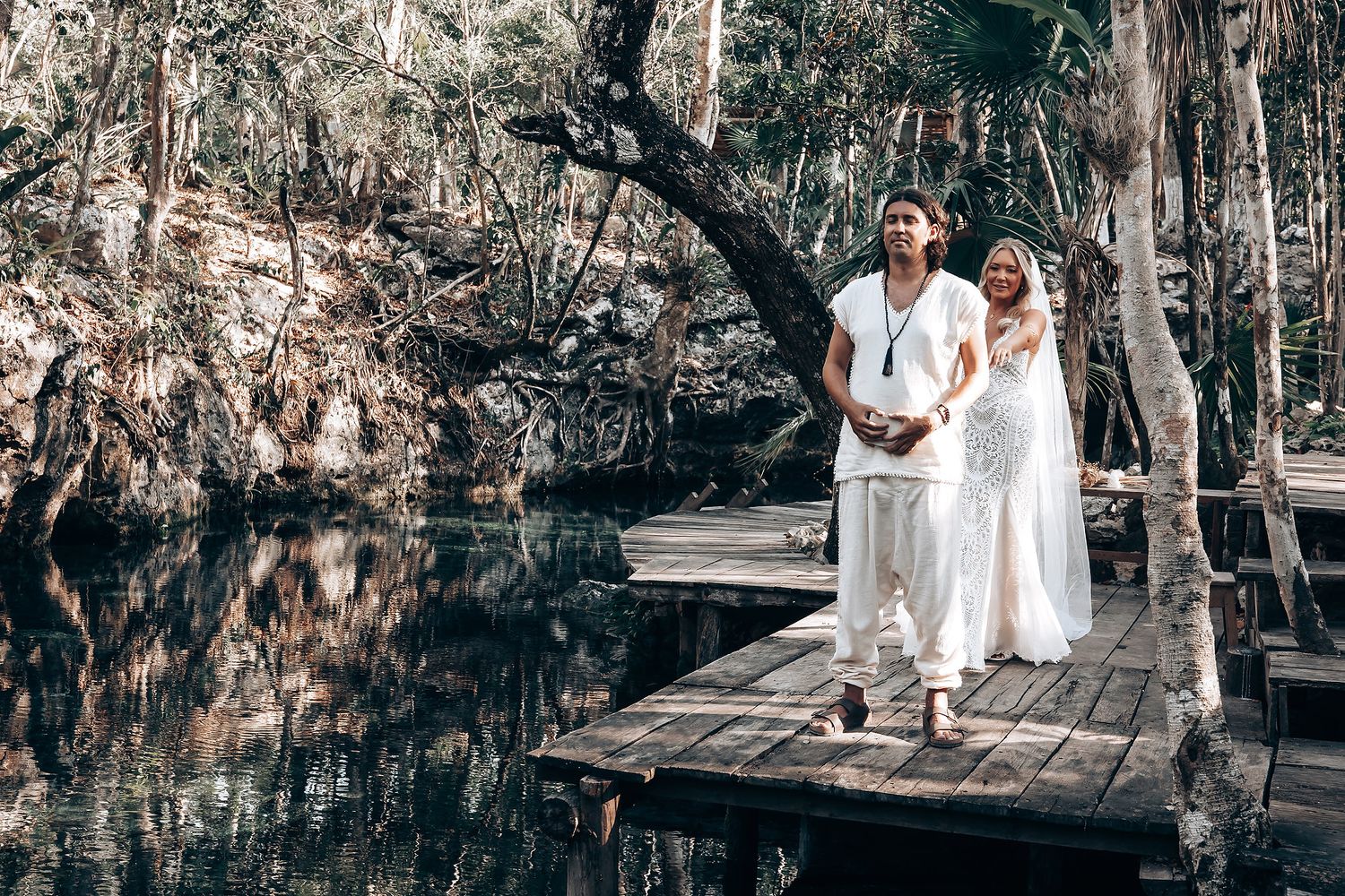 Couple in white attire poses together on a wooden boardwalk surrounded by lush trees and water.