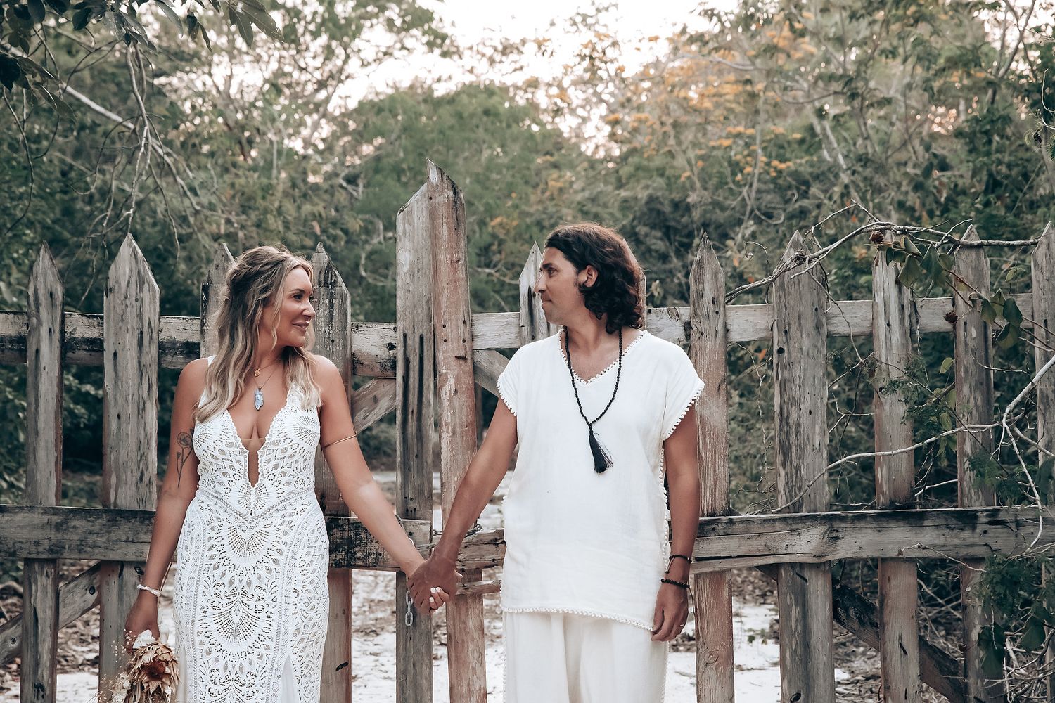 Two people in coordinating white ensembles walk hand-in-hand through a scenic wooded area.