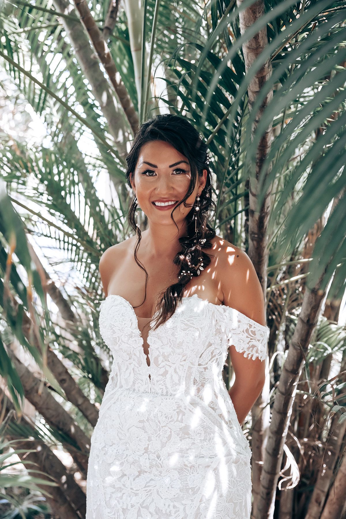 An elegant white lace wedding dress shimmers in dappled sunlight beneath tropical palm fronds.