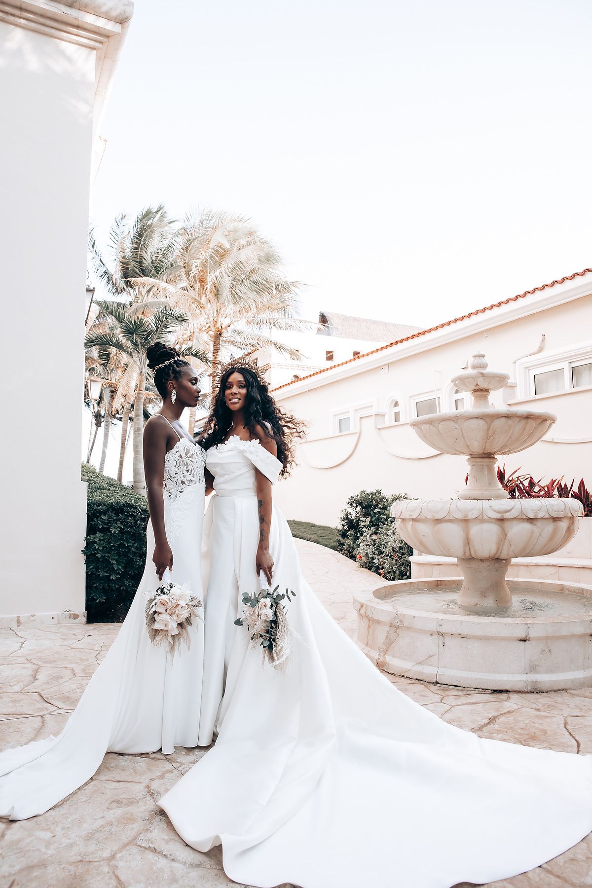 Bridal party in flowing white dresses posing near a Mediterranean-style fountain.