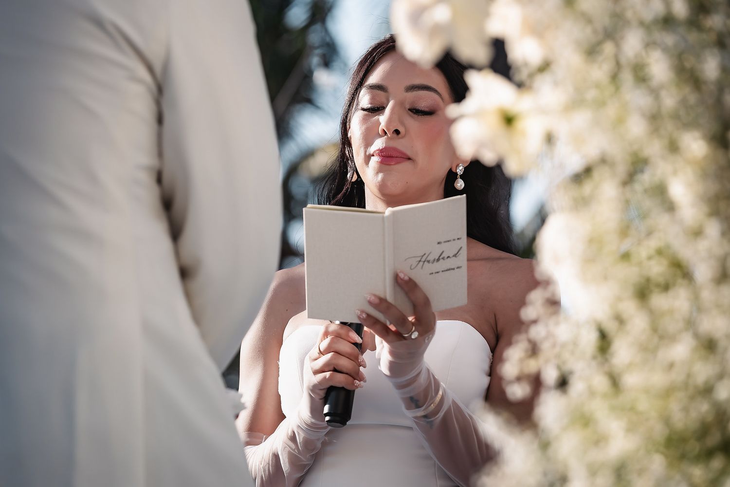 Bride reads vows from a small book while holding a microphone at an outdoor wedding ceremony.