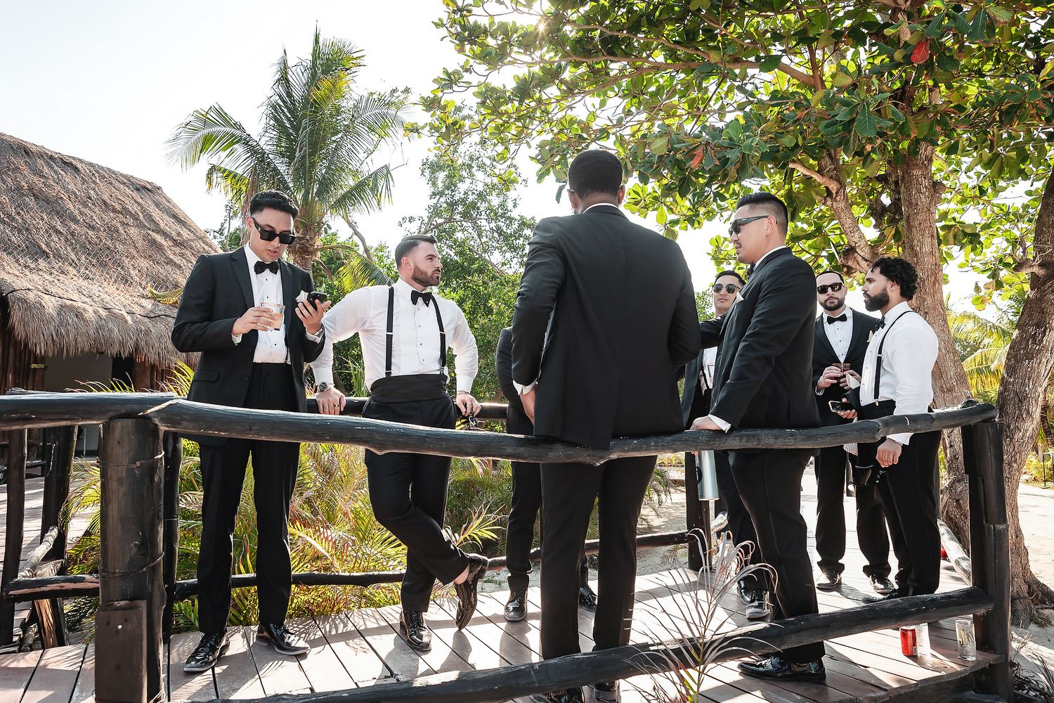 Groomsmen in black tuxedos socializing on a wooden deck at a tropical outdoor wedding venue.