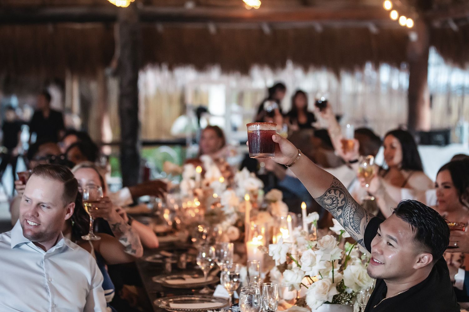 Guests raising glasses in a toast at an elegant candlelit wedding reception under a thatched roof venue.