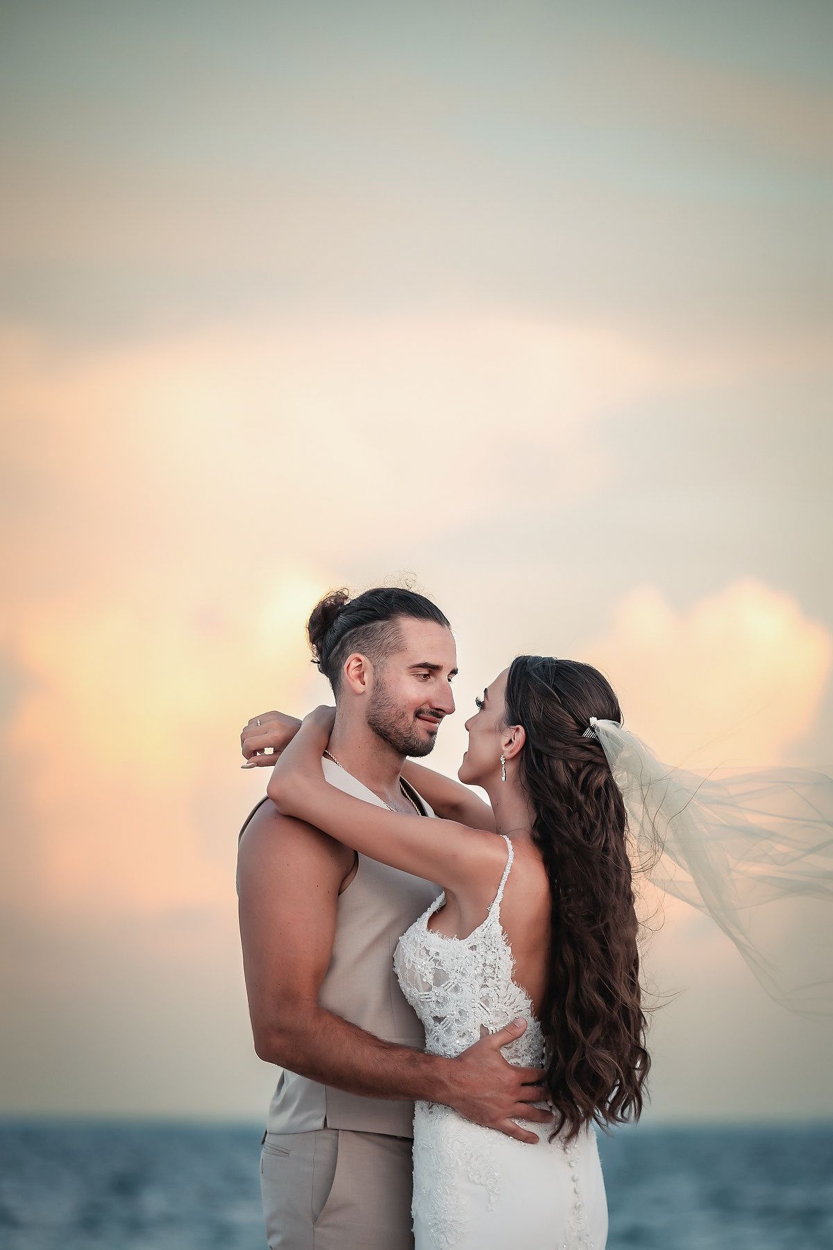 A couple in wedding attire embraces romantically on a beach at sunset, with the bride's veil flowing in the breeze.