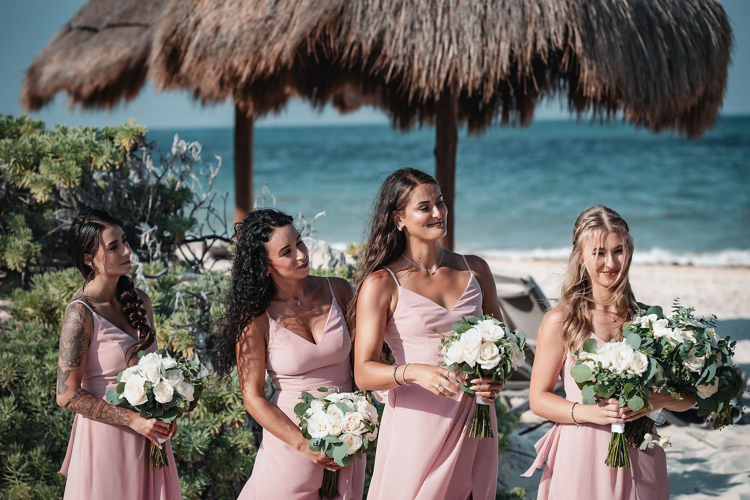 Four bridesmaids in pink dresses holding white bouquets on a tropical beach wedding with a thatched umbrella.