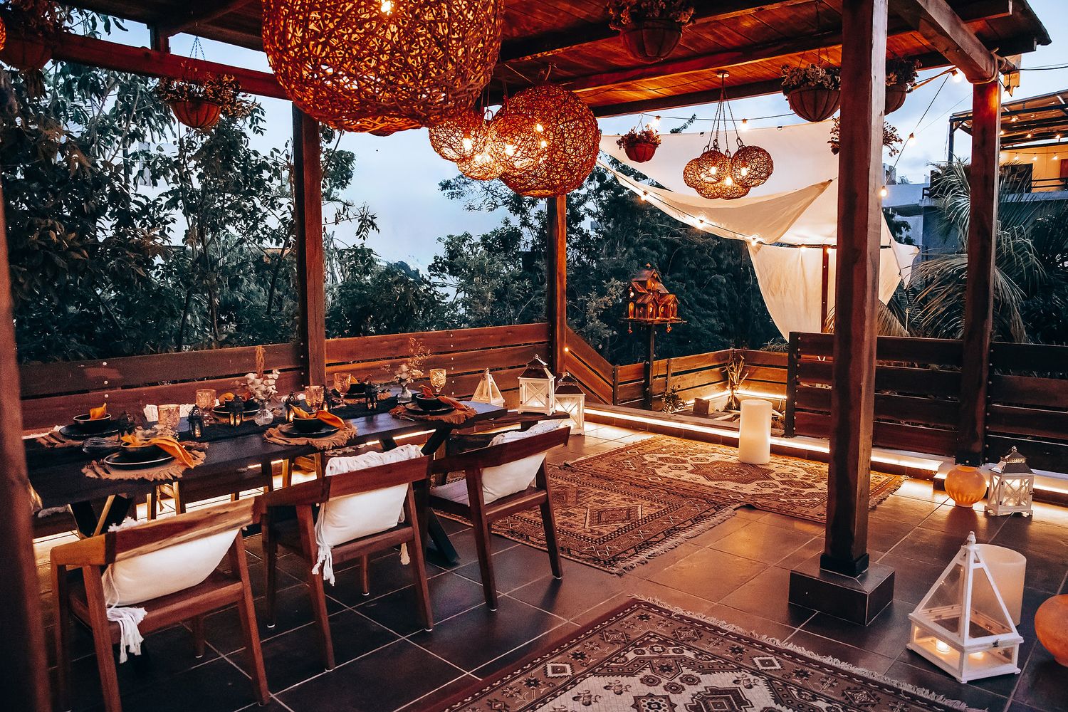 Elegant outdoor terrace dinner table set under warm wicker chandeliers with lush greenery at twilight.