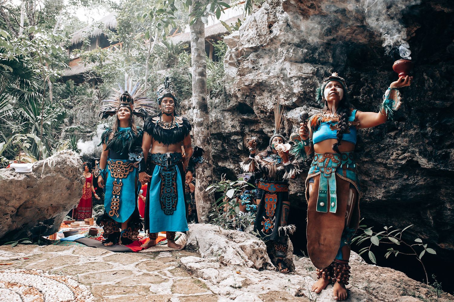 Three women in colorful Mayan warrior costumes perform a traditional ceremony among rocky jungle ruins.