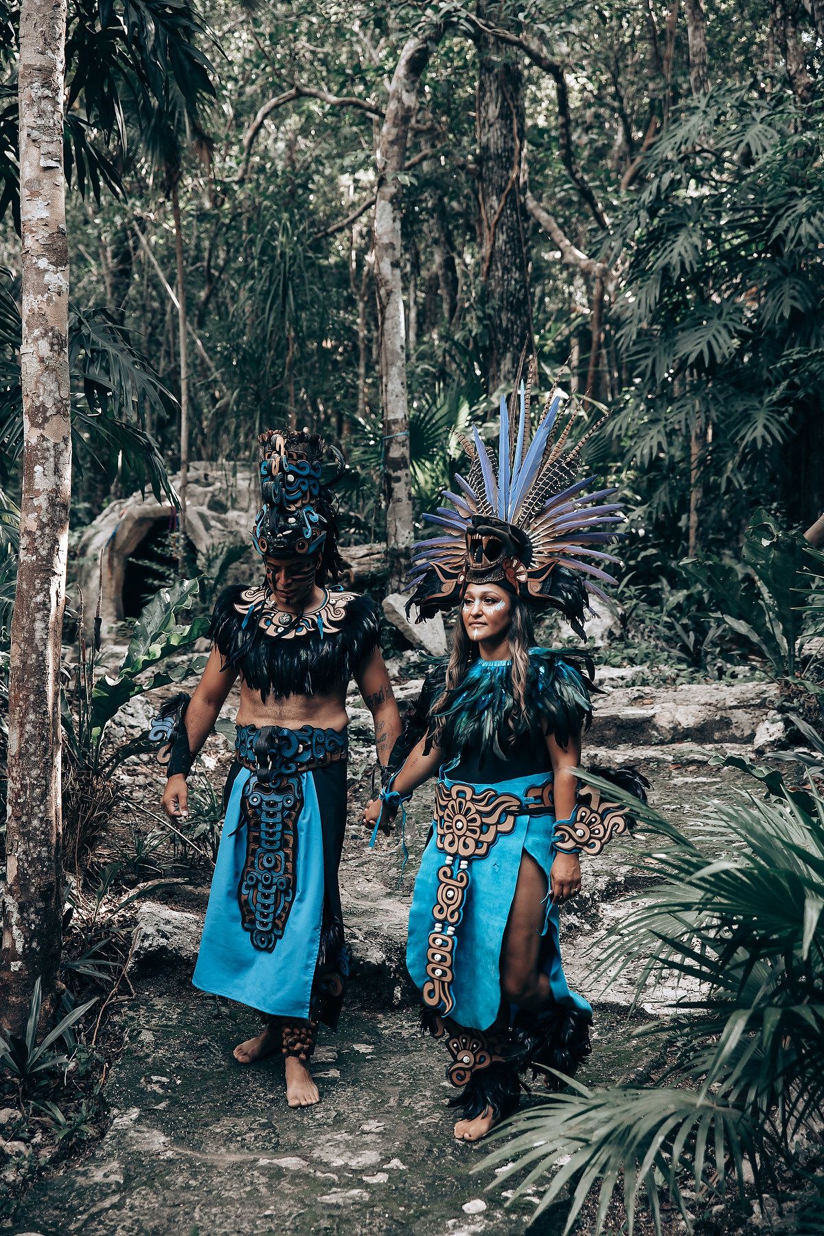 Two people in vibrant blue Mayan ceremonial costumes with feathered headdresses walking through a lush jungle.