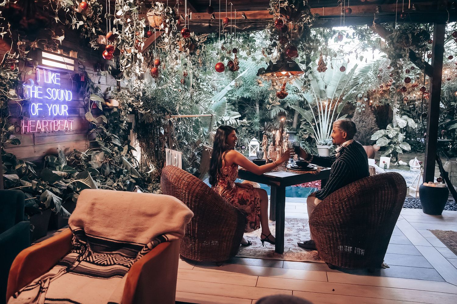 Couple dining at a romantic restaurant surrounded by lush greenery and festive holiday decorations with neon signage.