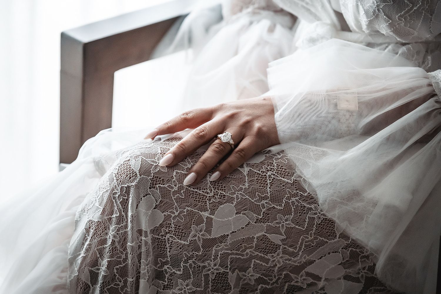 Close-up of a bride's hand with an elegant diamond ring resting on a lace wedding dress.