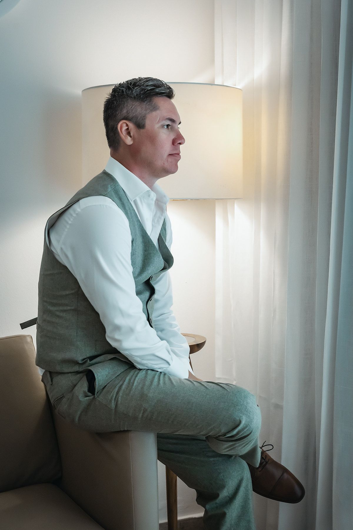 A well-dressed man in a green vest and trousers sits thoughtfully by a lamp near curtained windows.