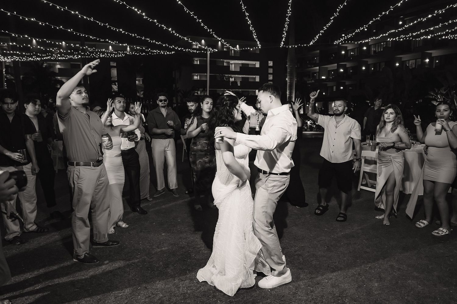 Bride and groom share their first dance surrounded by guests under twinkling string lights at outdoor wedding reception.