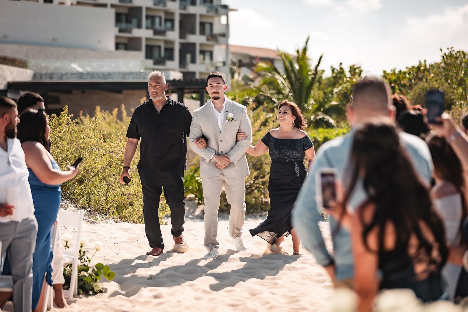 Groom walks down beach wedding aisle with parents on sunny day.