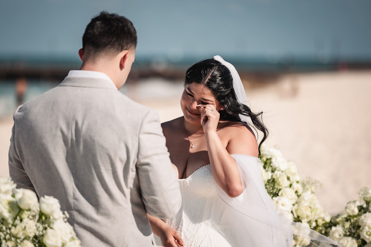 Emotional bride wiping tears during beach wedding ceremony with groom in gray suit, white floral arrangements.