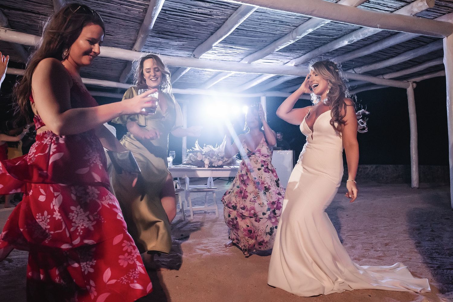 Bride in white gown dancing joyfully with guests in colorful dresses at an outdoor beach wedding reception.