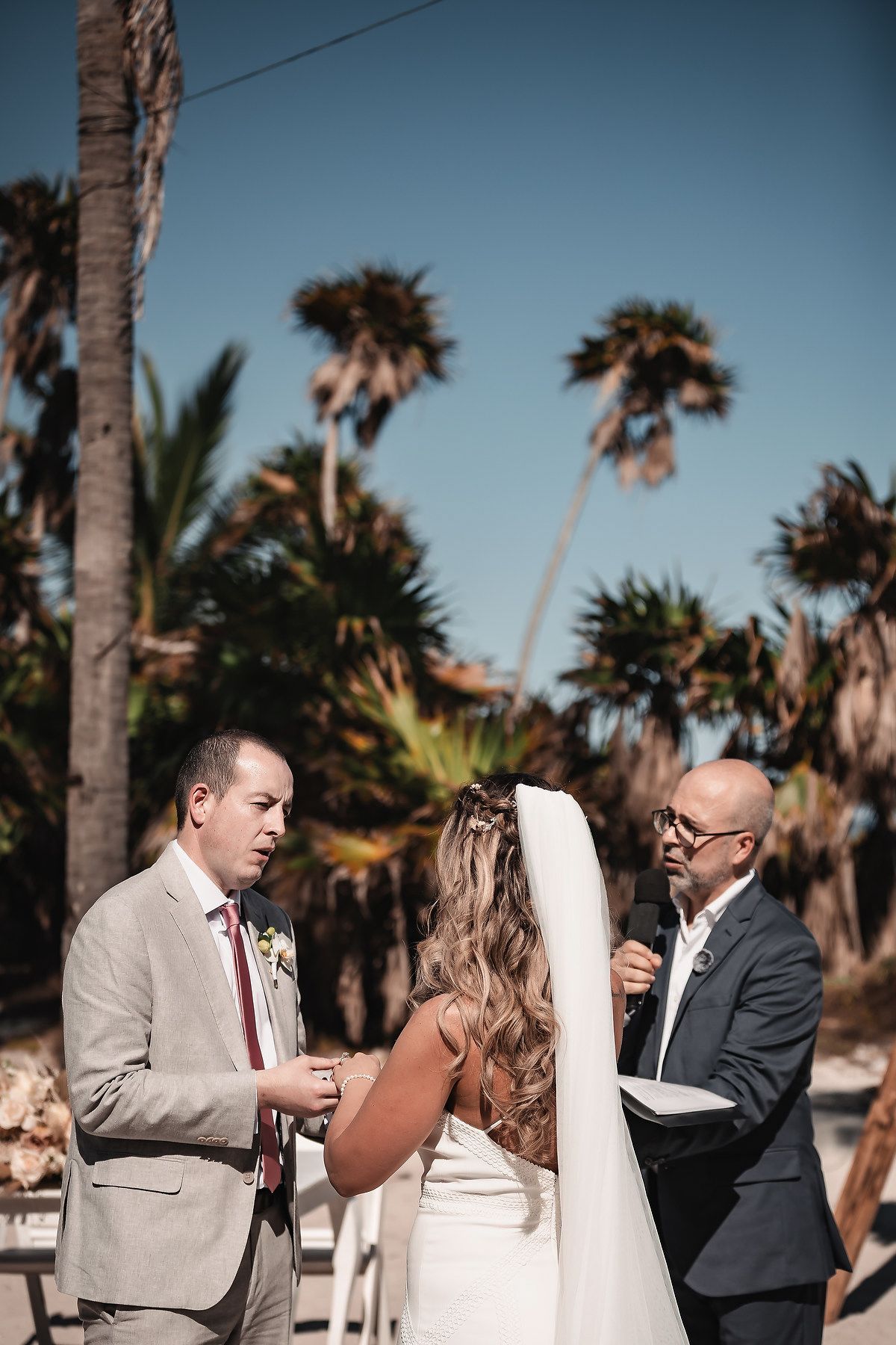 Outdoor beach wedding ceremony with groom exchanging vows, bride in white veil, officiant holding mic, palm trees under blue sky.