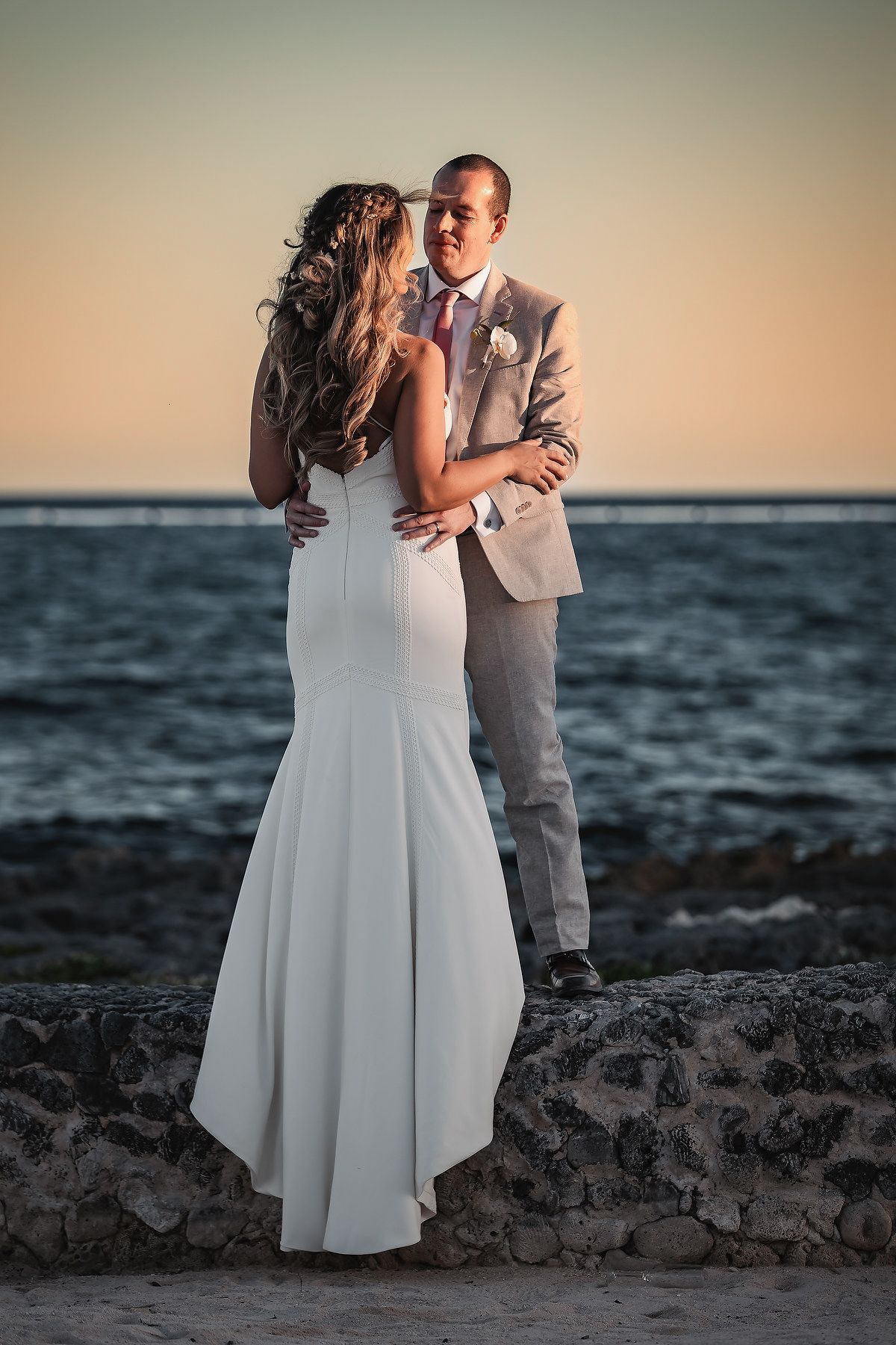 Bride and groom embrace at sunset by the ocean, bride in white gown, groom in tan suit with boutonniere.