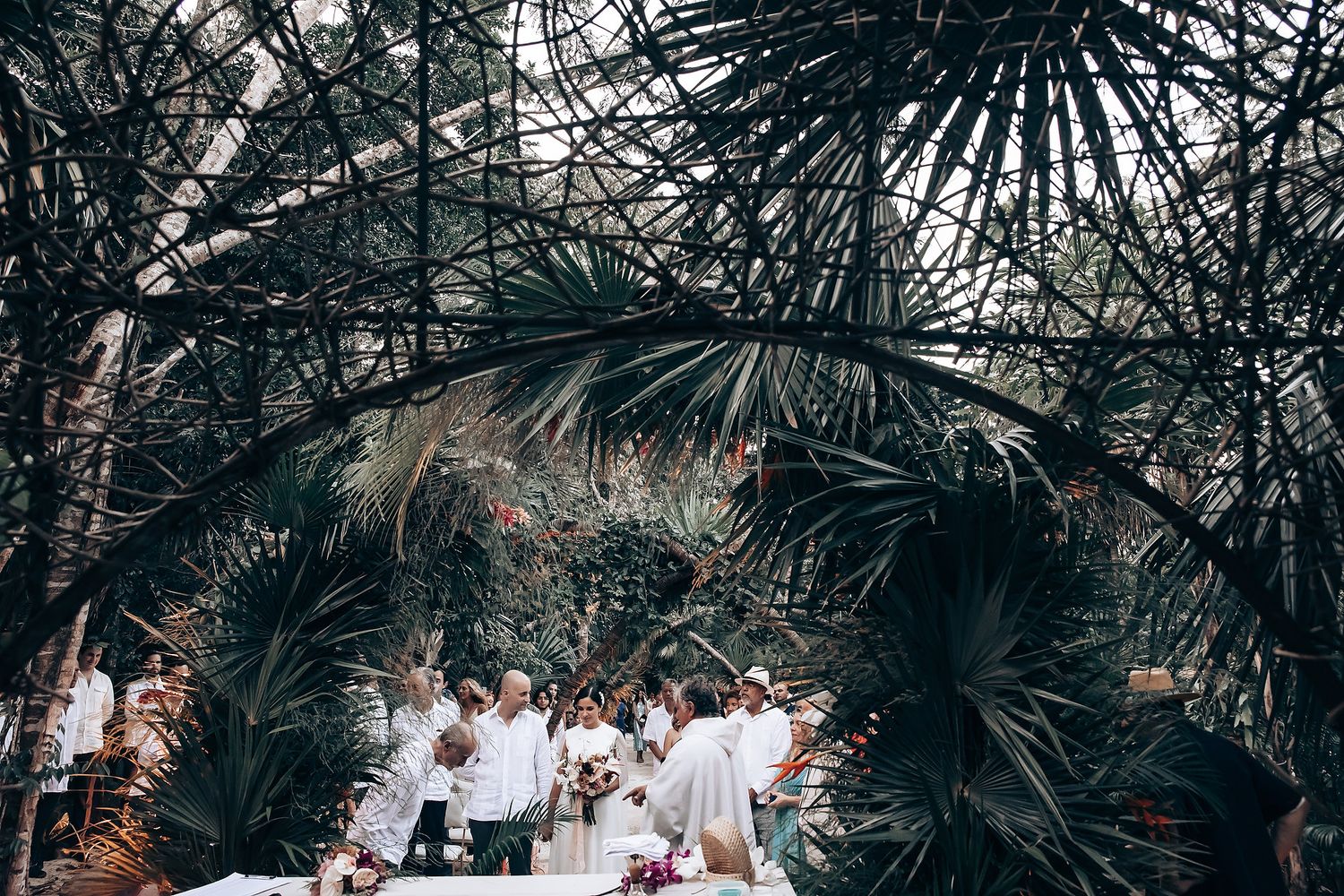 A tropical wedding ceremony setup under lush palm fronds with guests gathered in white attire.