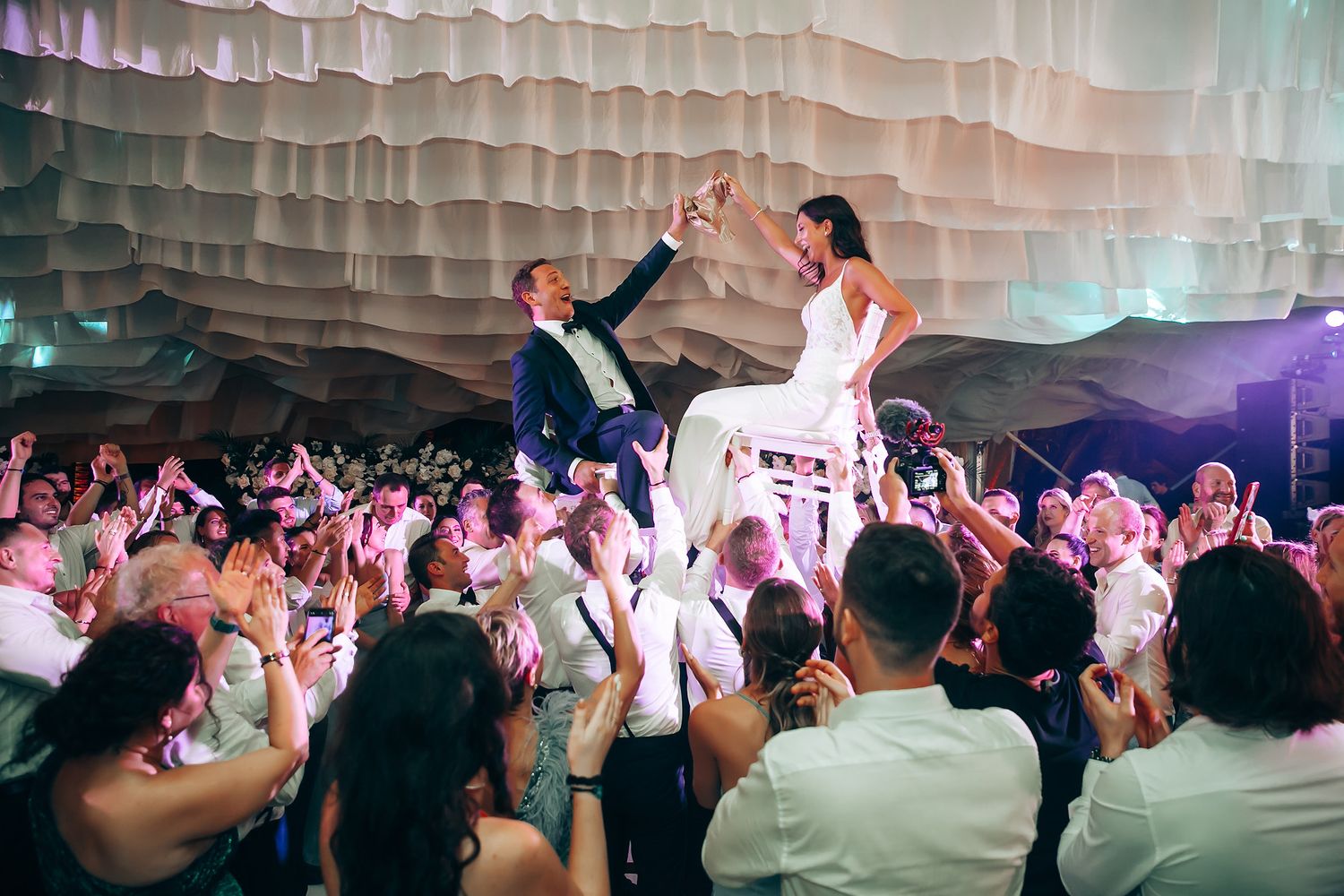 Wedding reception dance floor under white draped ceiling with colorful lighting.