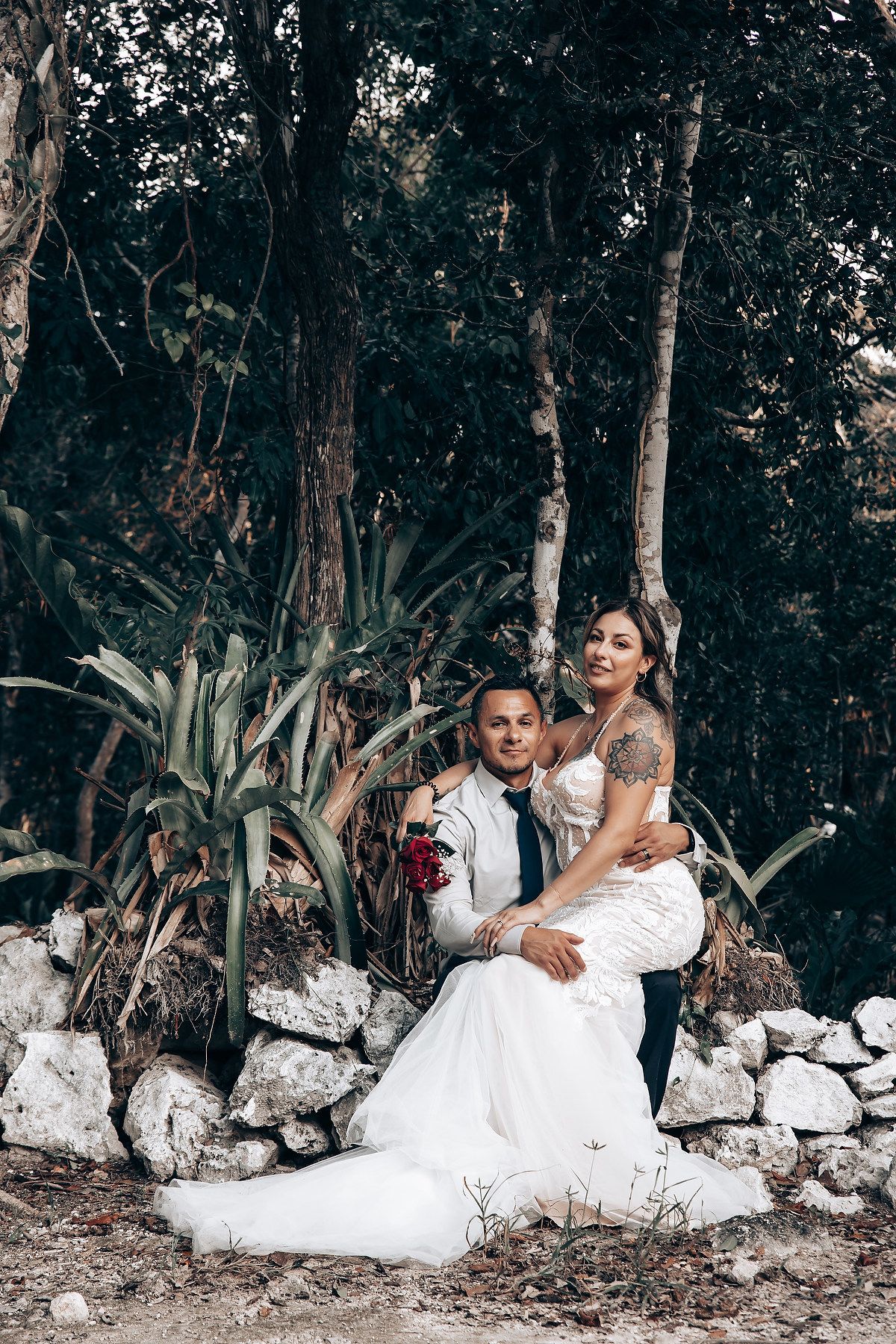 A couple in wedding attire poses against a rustic stone wall surrounded by tropical plants.