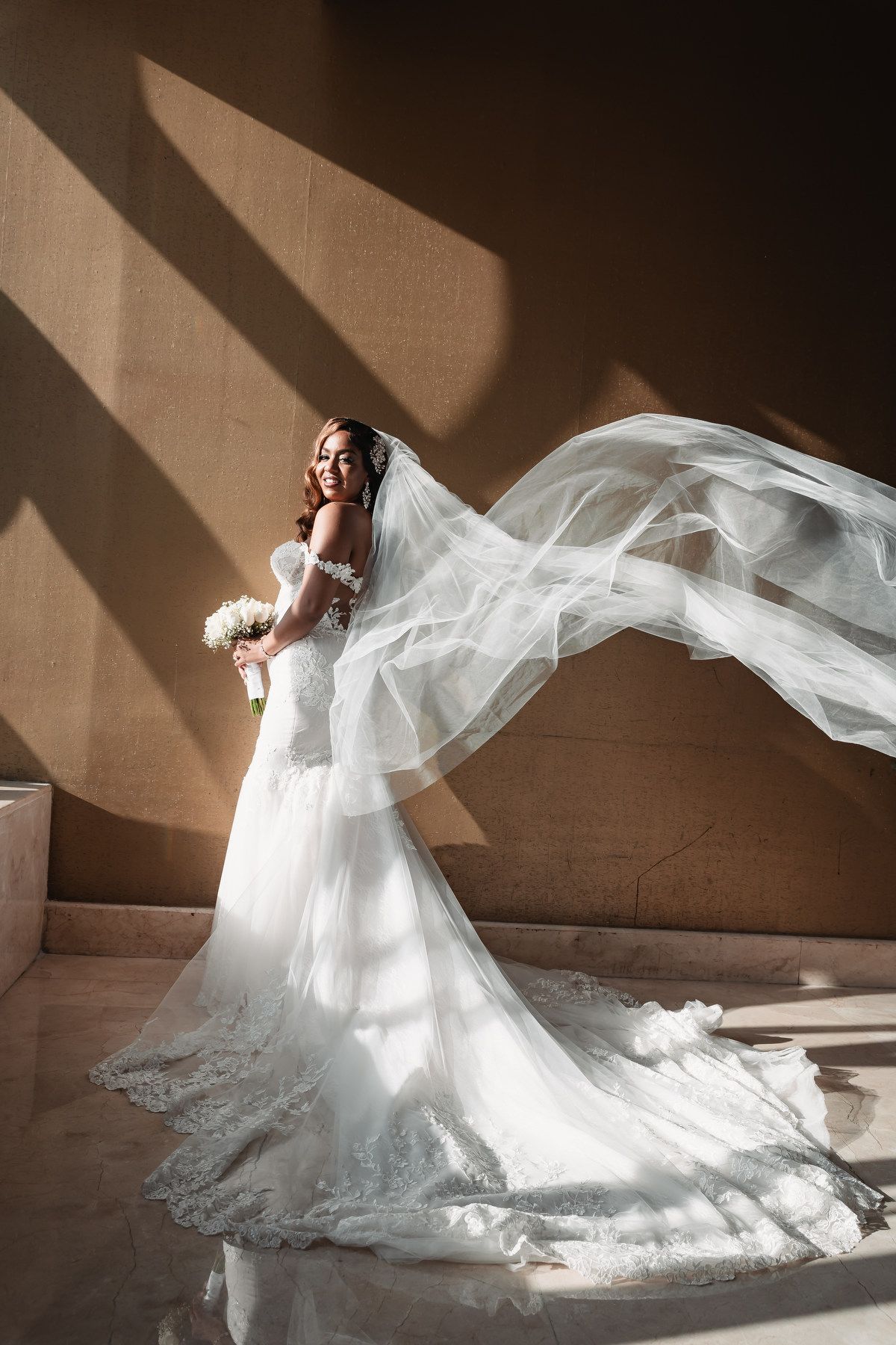 Stunning bride in lace mermaid gown with flowing veil caught in sunlight against a warm stone wall.