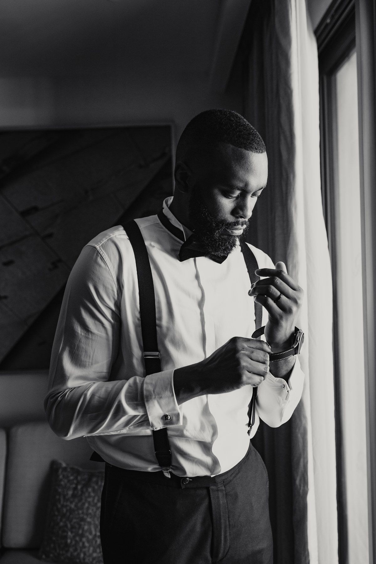 Black and white portrait of groom in suspenders and bow tie by window light.