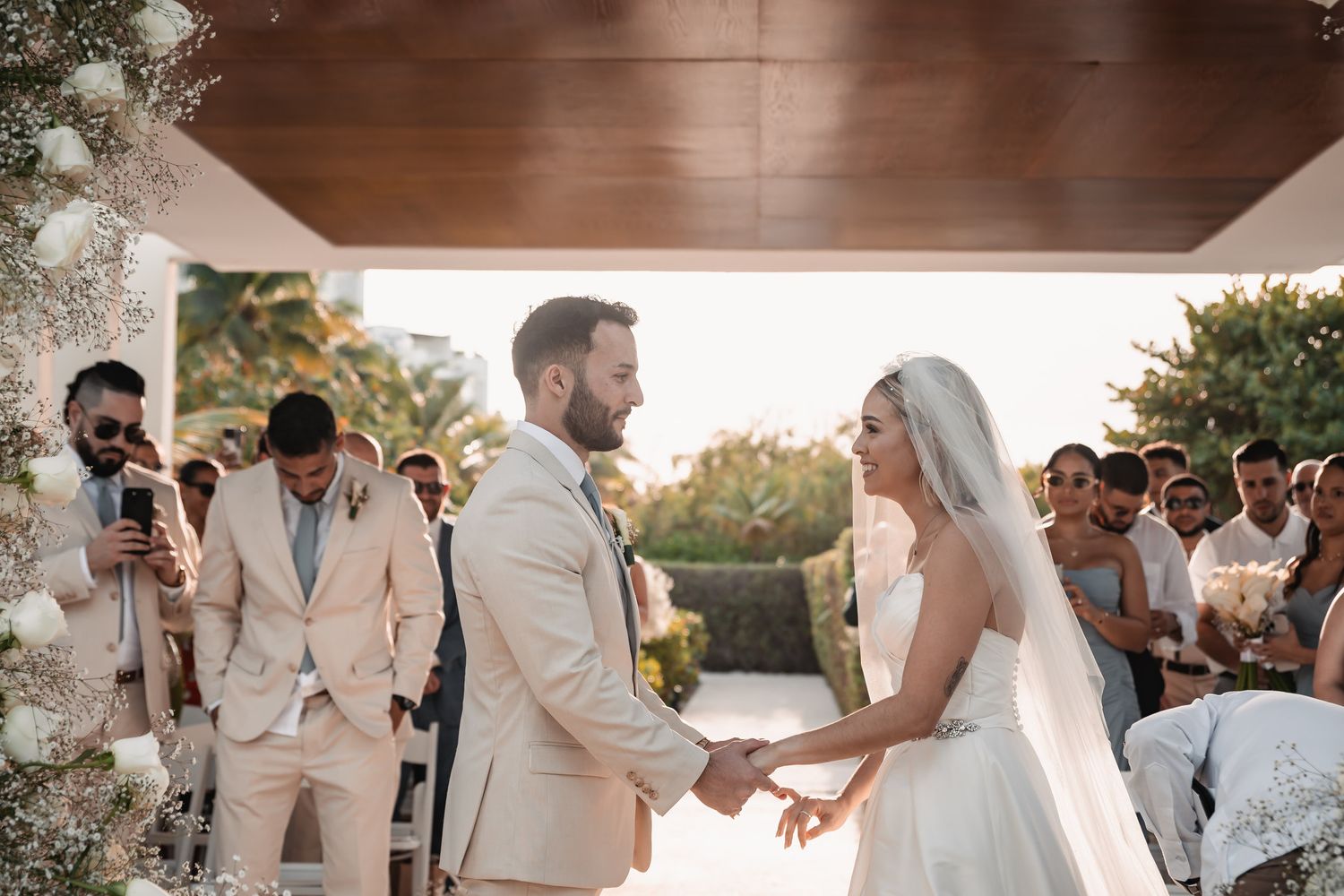 Bride and groom hold hands at outdoor wedding ceremony with guests seated behind them.