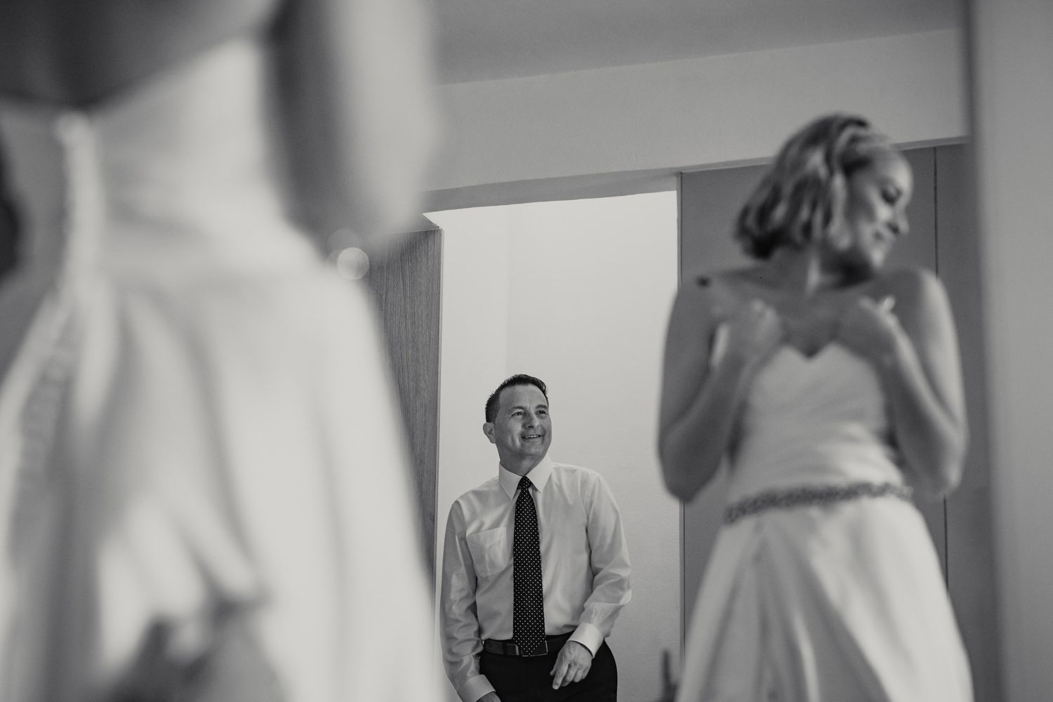 Black and white photo of groom watching bride get ready in mirror reflection during wedding preparations.