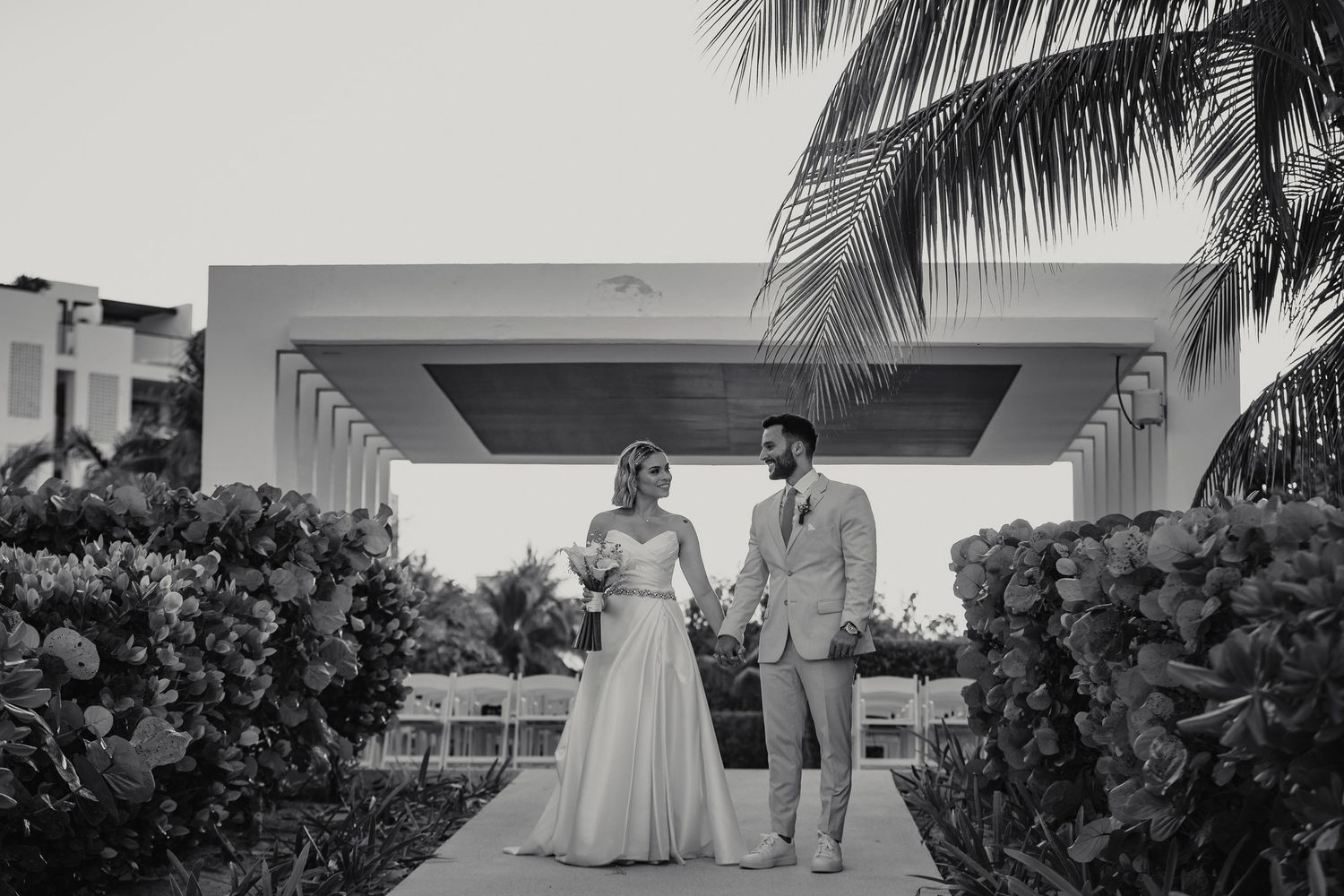 Black and white photo of bride and groom standing under modern pergola at tropical venue.