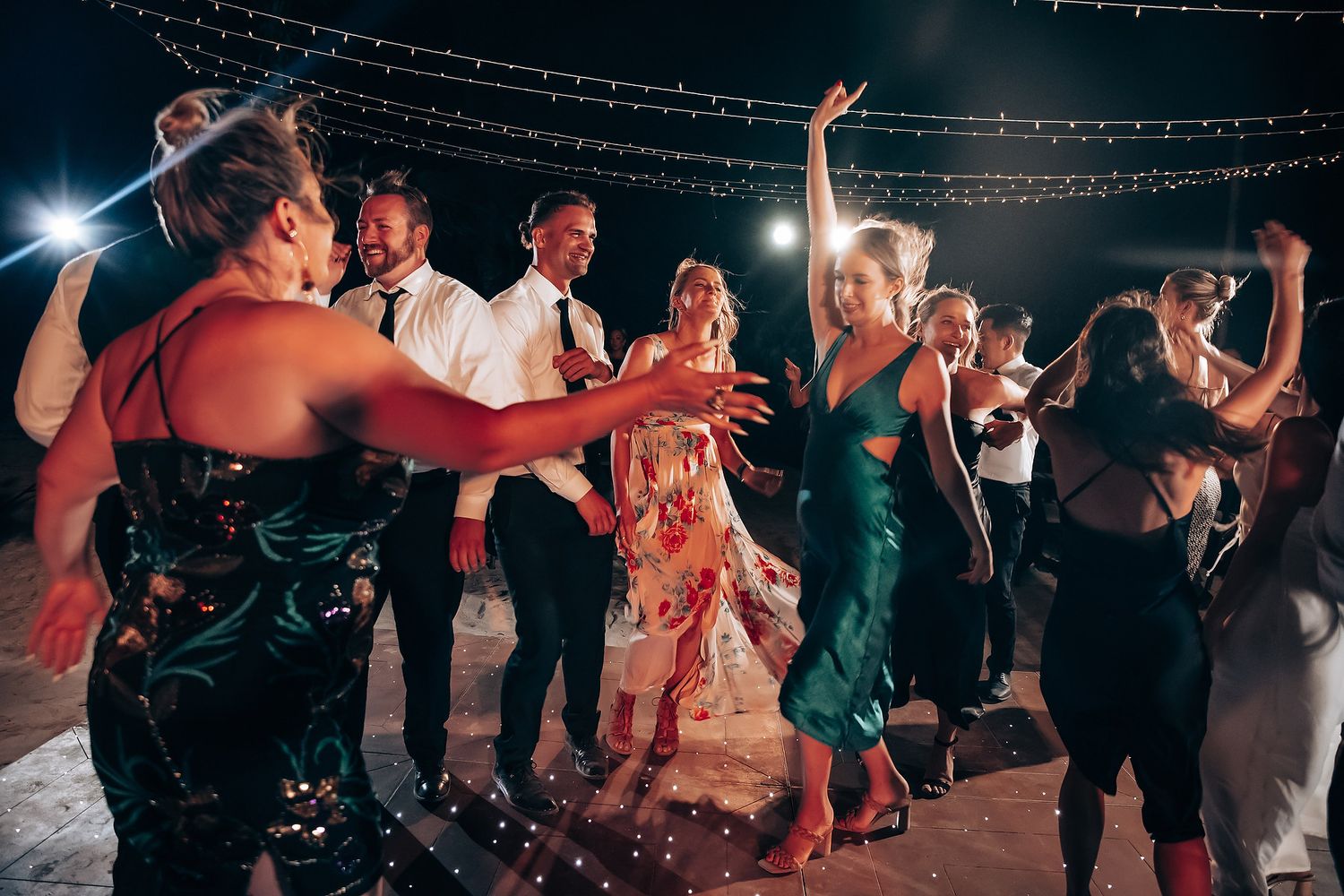 Wedding party dances under twinkling string lights at an outdoor evening reception.