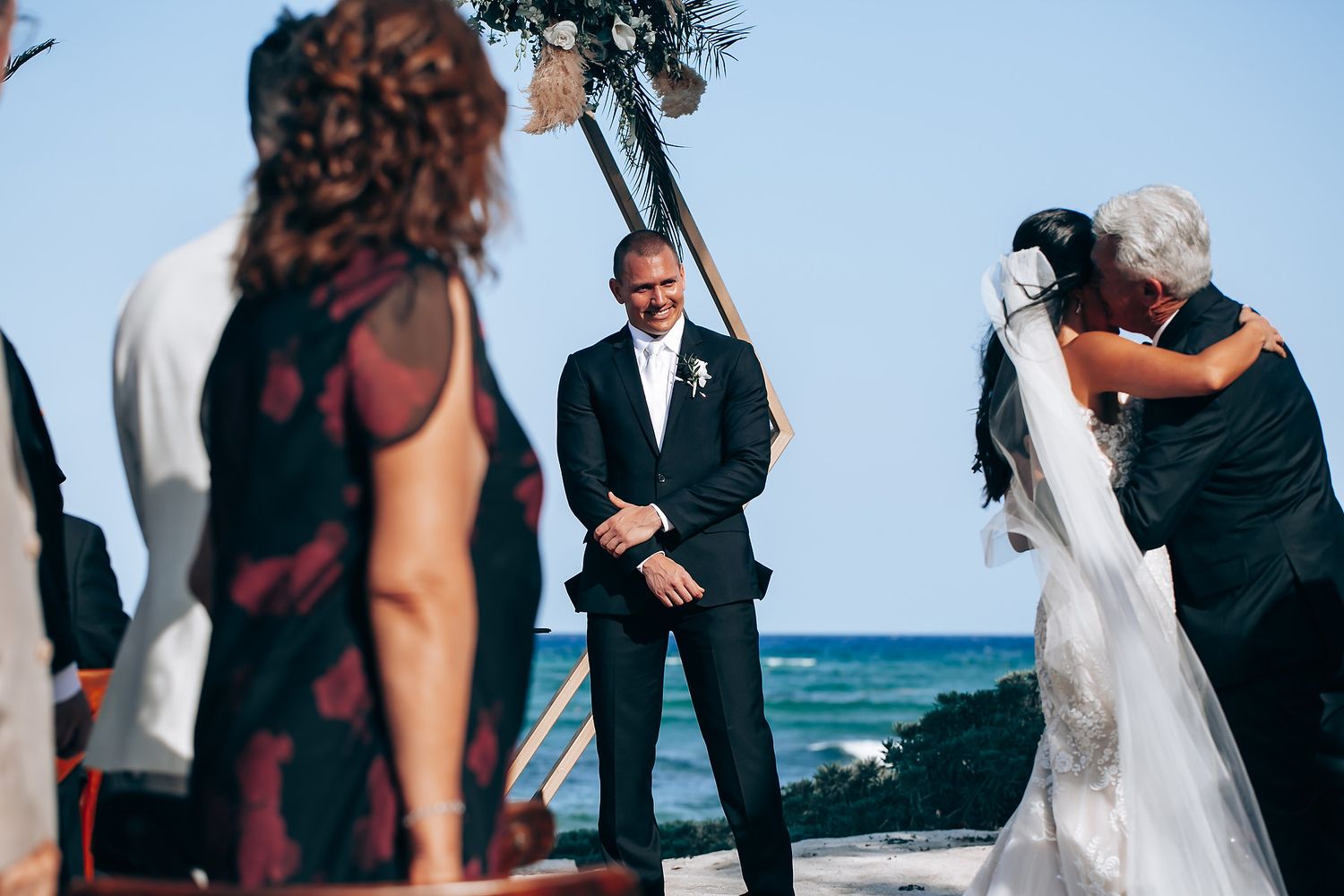 A beachside wedding ceremony unfolds with attendees gathered under blue skies near the ocean.