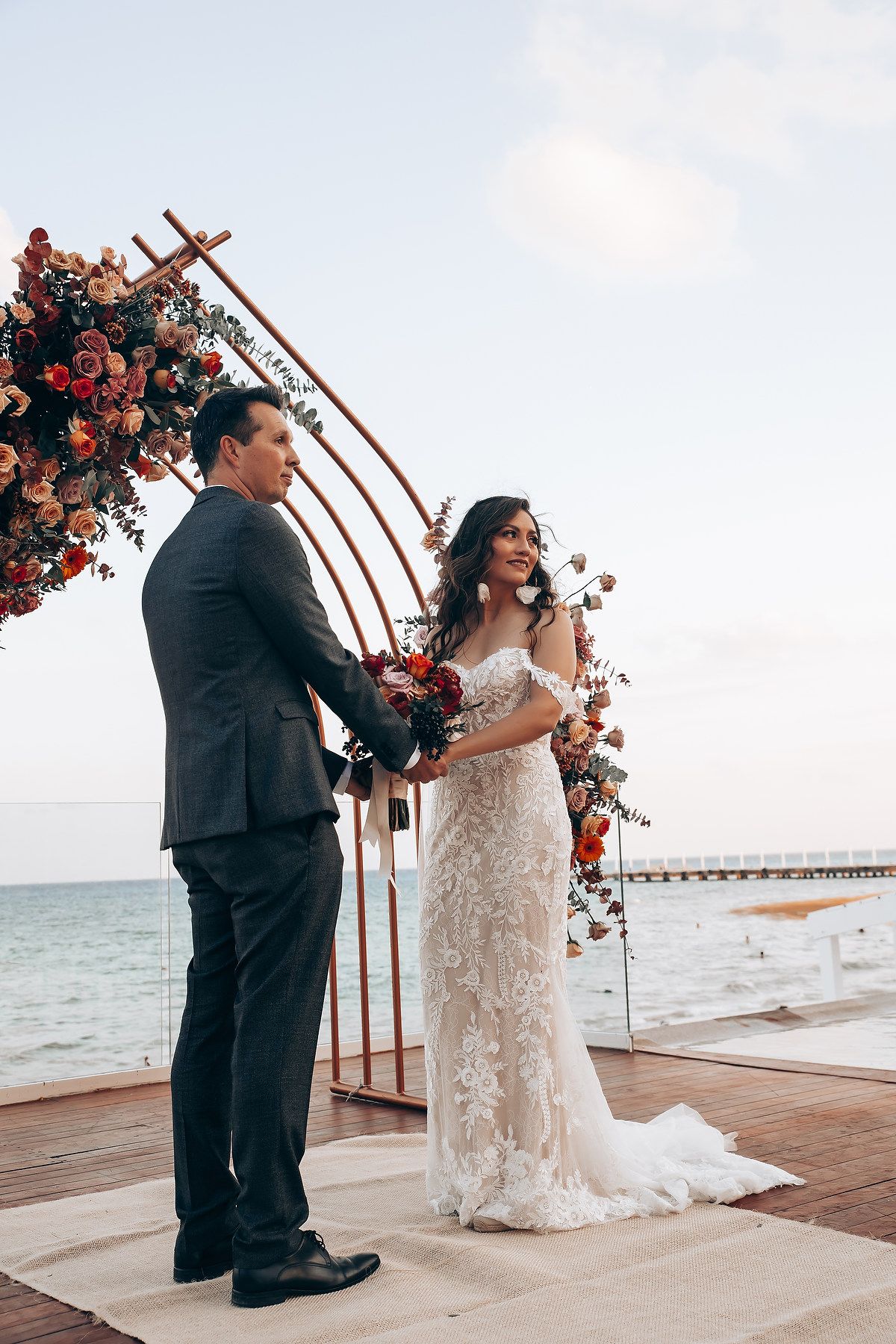 A wedding ceremony on a seaside deck with an ornate floral arch featuring coral and pink roses.