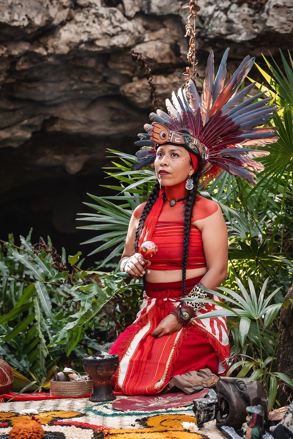 Woman in traditional Mayan red costume and feathered headdress kneeling outdoors in a lush jungle setting.