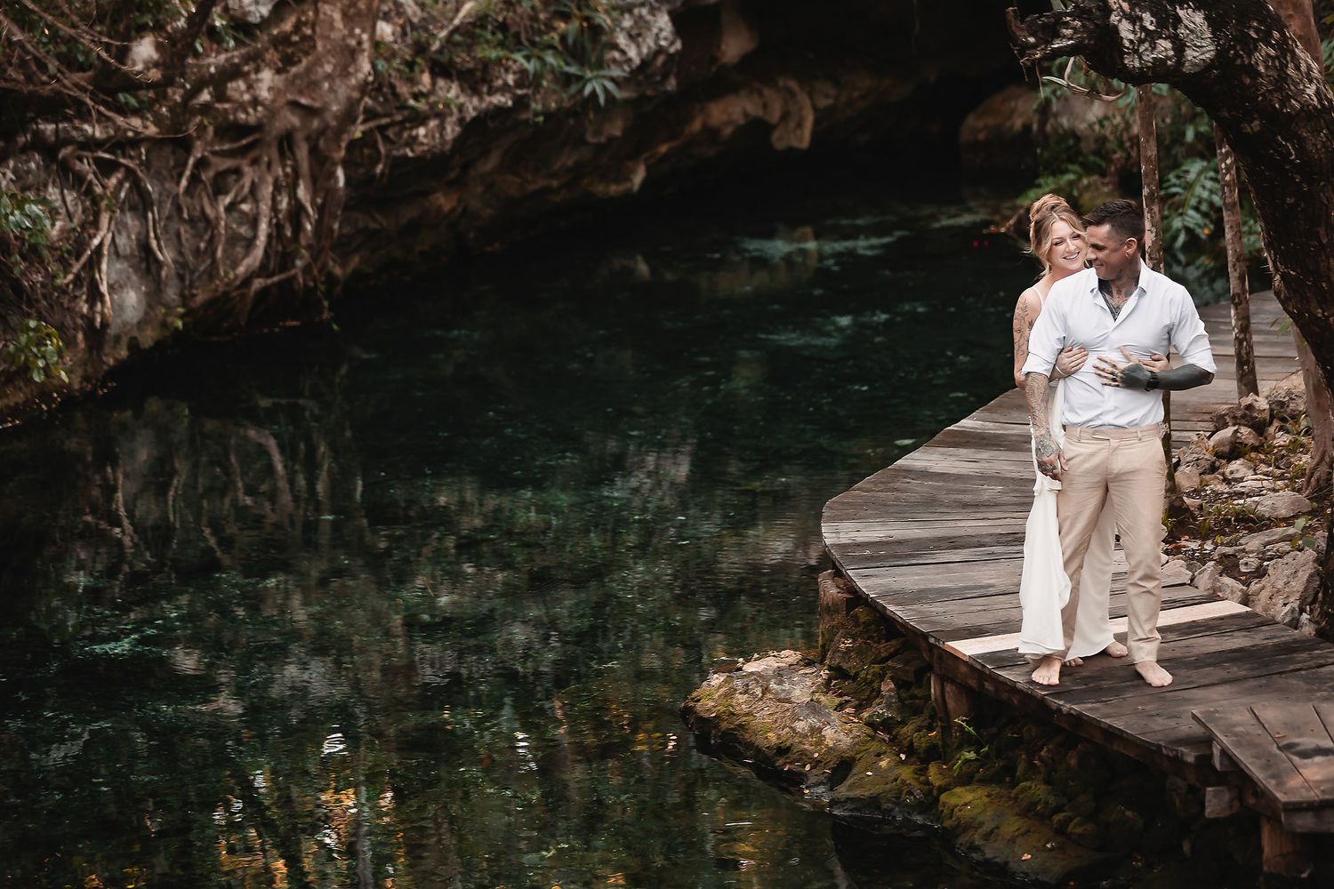Couple standing on a wooden dock over crystal clear cenote waters surrounded by lush tropical greenery.