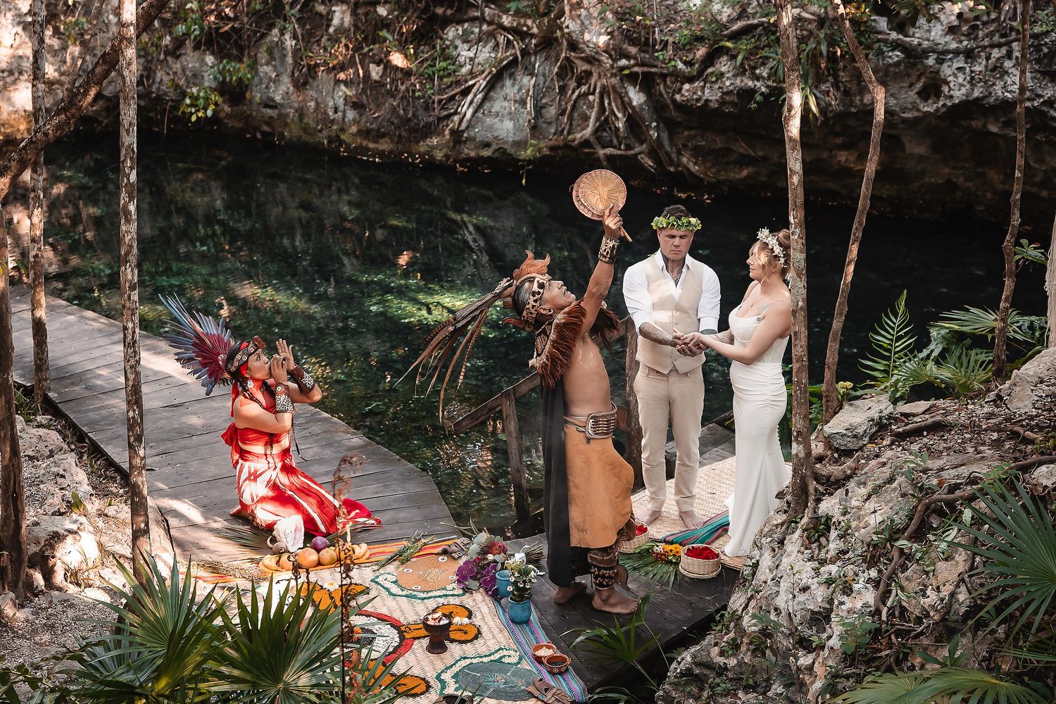 Couple stands at jungle cenote altar during Mayan wedding ceremony with costumed officiants and colorful decor.