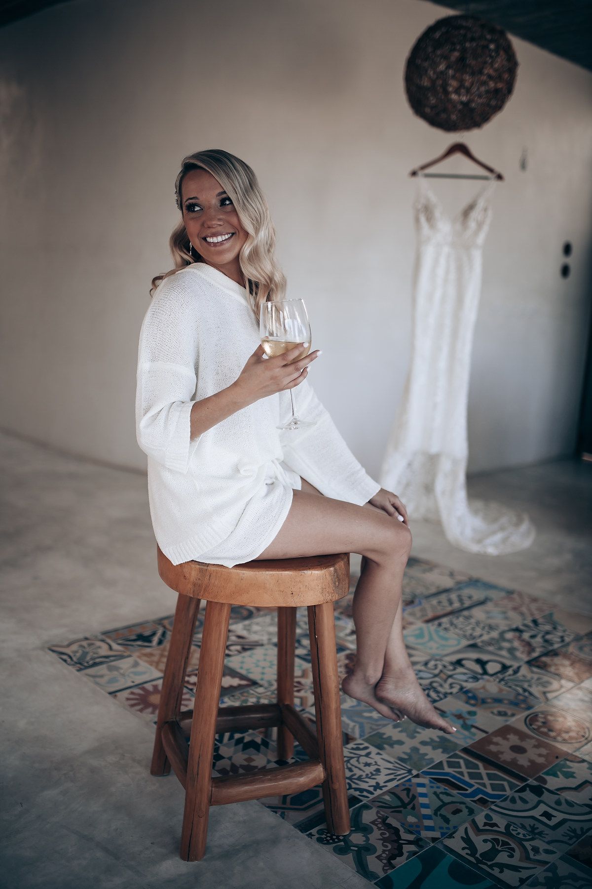A woman in a white robe sits on a wooden stool with a wedding dress hanging in the background.